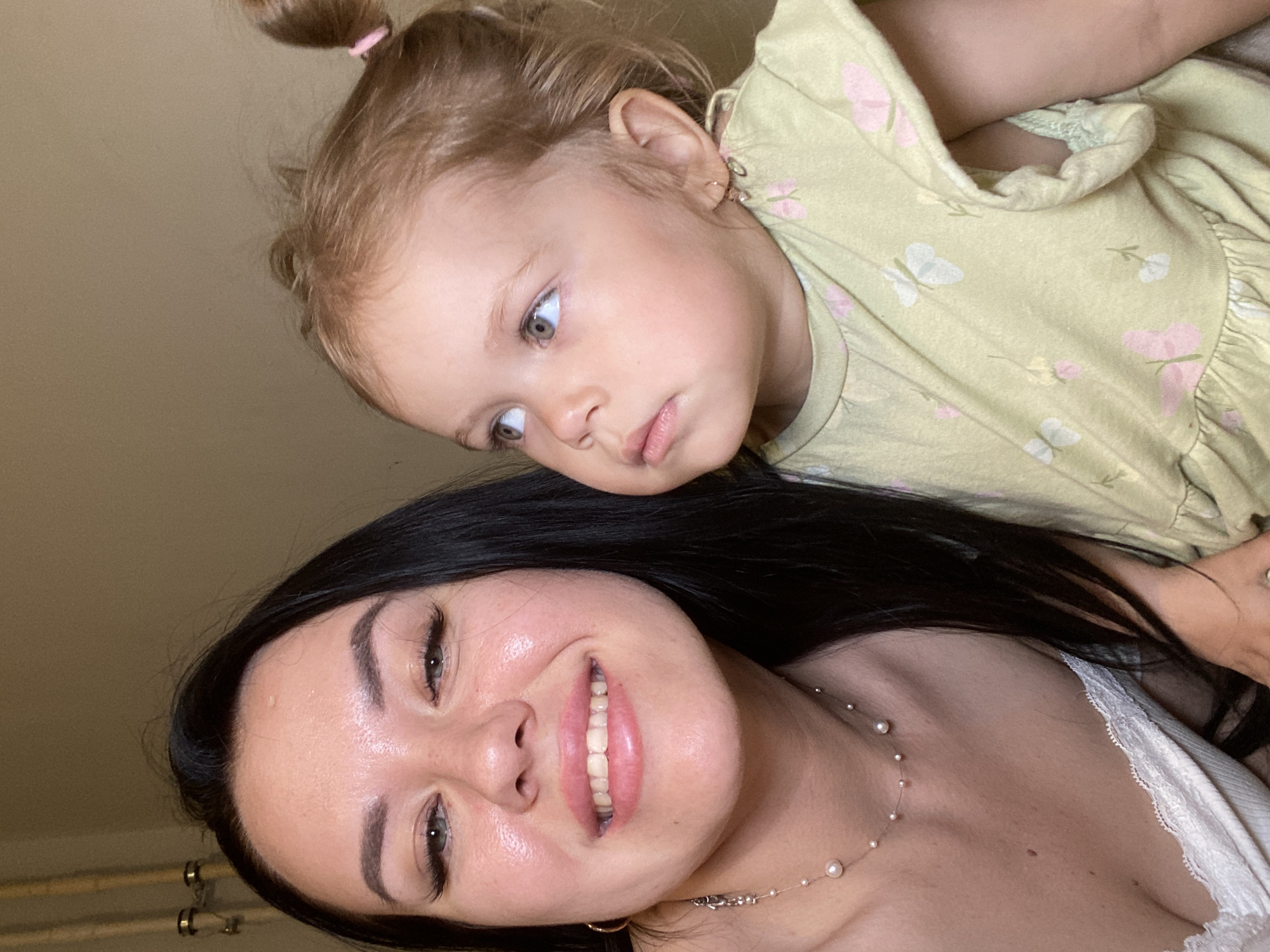 An undated, medium shot selfie shows a woman and a young child indoors. The woman, on the left, has long black hair and is wearing a white top with lace trim and a pearl necklace. She is smiling and looking at the camera. The child, on the right, has blonde hair in two buns and is wearing a light green dress with a butterfly pattern. The child is looking upwards and to the right. The background appears to be a plain, light-colored wall with some vertical pipes visible on the left. No specific location in Chaikovsky, Russia, can be identified from the image.