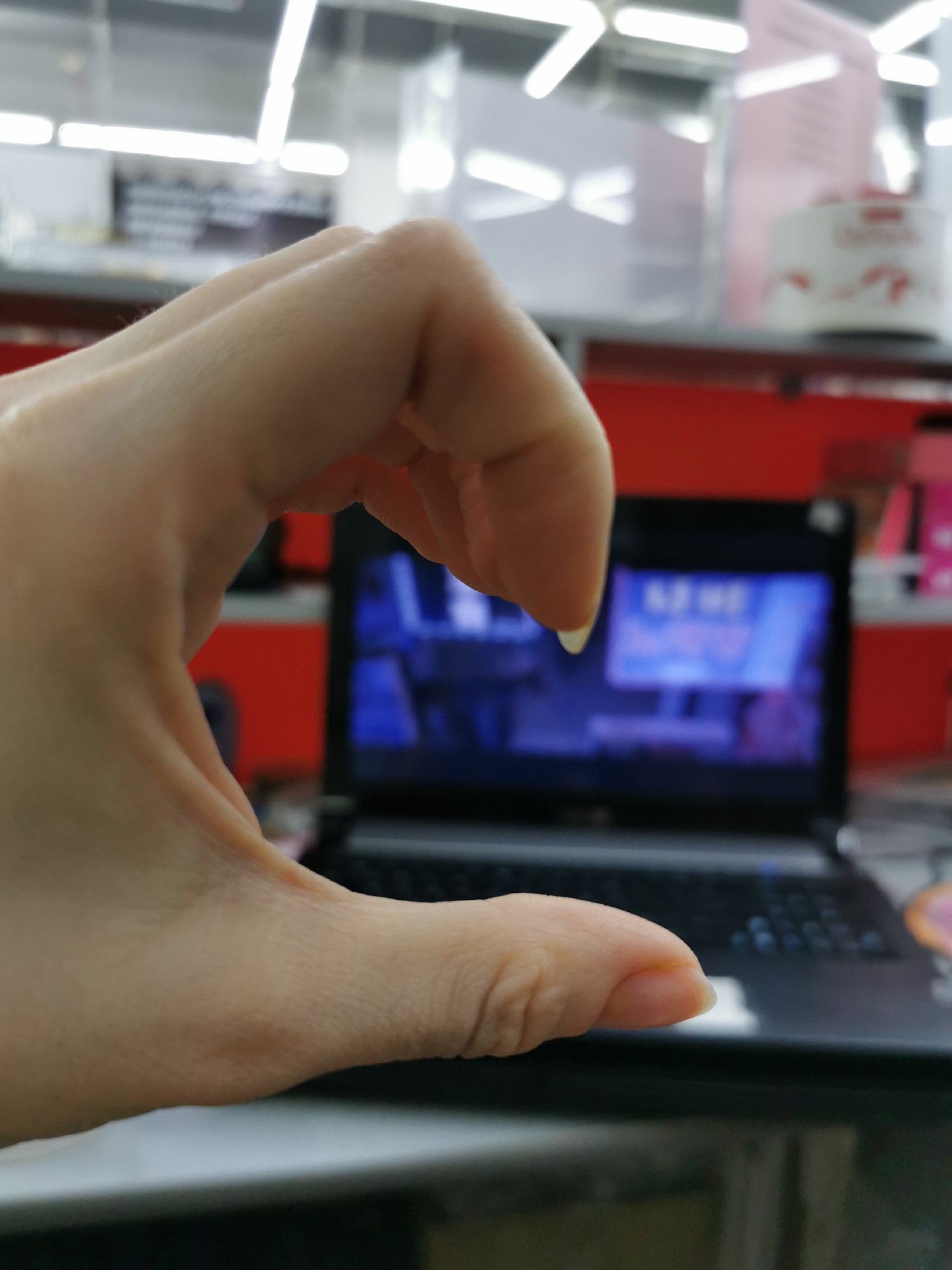 A human hand is positioned in the foreground, partially obscuring a laptop. The hand is pale, with the fingers curved into a C-shape. The laptop is open and displaying a screen with indistinct content. The background is blurry, suggesting an indoor environment with shelves or displays in shades of red, white and black. Based on the visible elements, the location is a retail establishment.