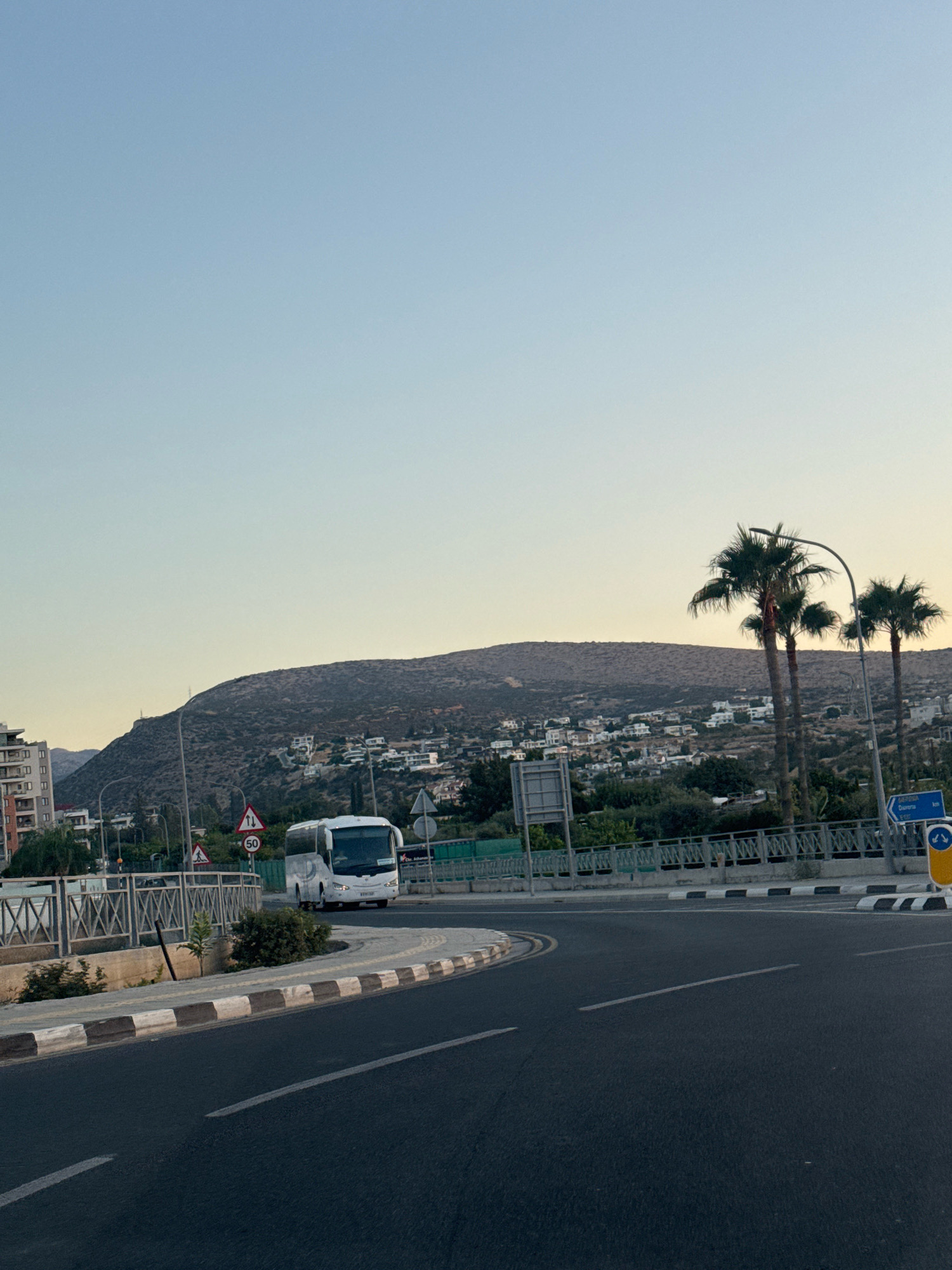 The image captures a view of a road intersection in Germasogeia Municipality, Cyprus, during what appears to be late afternoon or early evening, given the soft, warm light. A white bus is positioned at the intersection, suggesting it is either waiting to proceed or in the process of turning. The road surface is asphalt, with visible lane markings. A concrete barrier with alternating black and white painted sections separates the road from a sidewalk and a decorative metal fence.
In the background, a hillside is dotted with white buildings, indicating a residential area. Several palm trees are visible near the right side of the frame, characteristic of the Mediterranean climate. Traffic signs are present, including a speed limit sign indicating "50" and directional signs pointing towards "Limassol," "Diarizos," and "Erimi." A modern building, likely an apartment complex, stands on the left side of the image. The sky is clear and exhibits a gradient of pale blue to a lighter hue near the horizon.