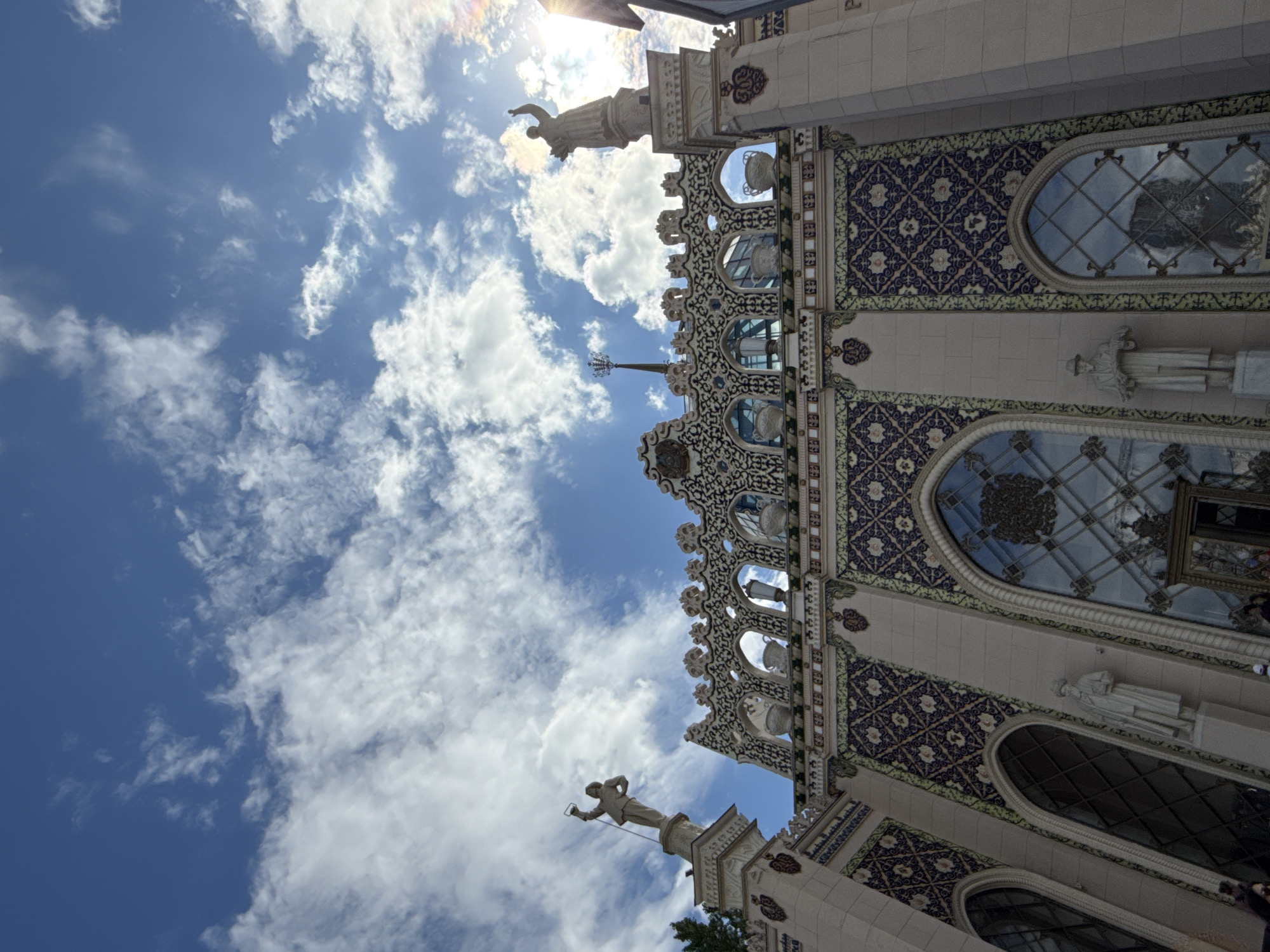 The image shows a low-angle view of the exterior of a building with ornate architectural details against a bright blue sky with fluffy white clouds. The building's facade is decorated with intricate patterns of blue and white tiles, as well as light-colored stone ornamentation. Several statues of human figures are visible on the building, perched on pedestals or integrated into the facade. Large arched windows with decorative latticework are also prominent features. The sun is shining brightly, casting a strong glare and creating a lens flare effect in the upper part of the image, suggesting it is daytime, likely mid-morning or afternoon. The overall impression is one of grandeur and historical significance. The scene appears to be set in an urban environment, given the architectural style and the presence of a building. While there are no explicit text elements visible, the architecture itself suggests a location with a rich history and possibly a cultural or artistic purpose. The style of the building, with its detailed tile work and classical statues, could be indicative of a museum, a historical landmark, or a public institution in Moscow, Russia. There is no discernible activity or event taking place, other than the static presentation of the architecture.