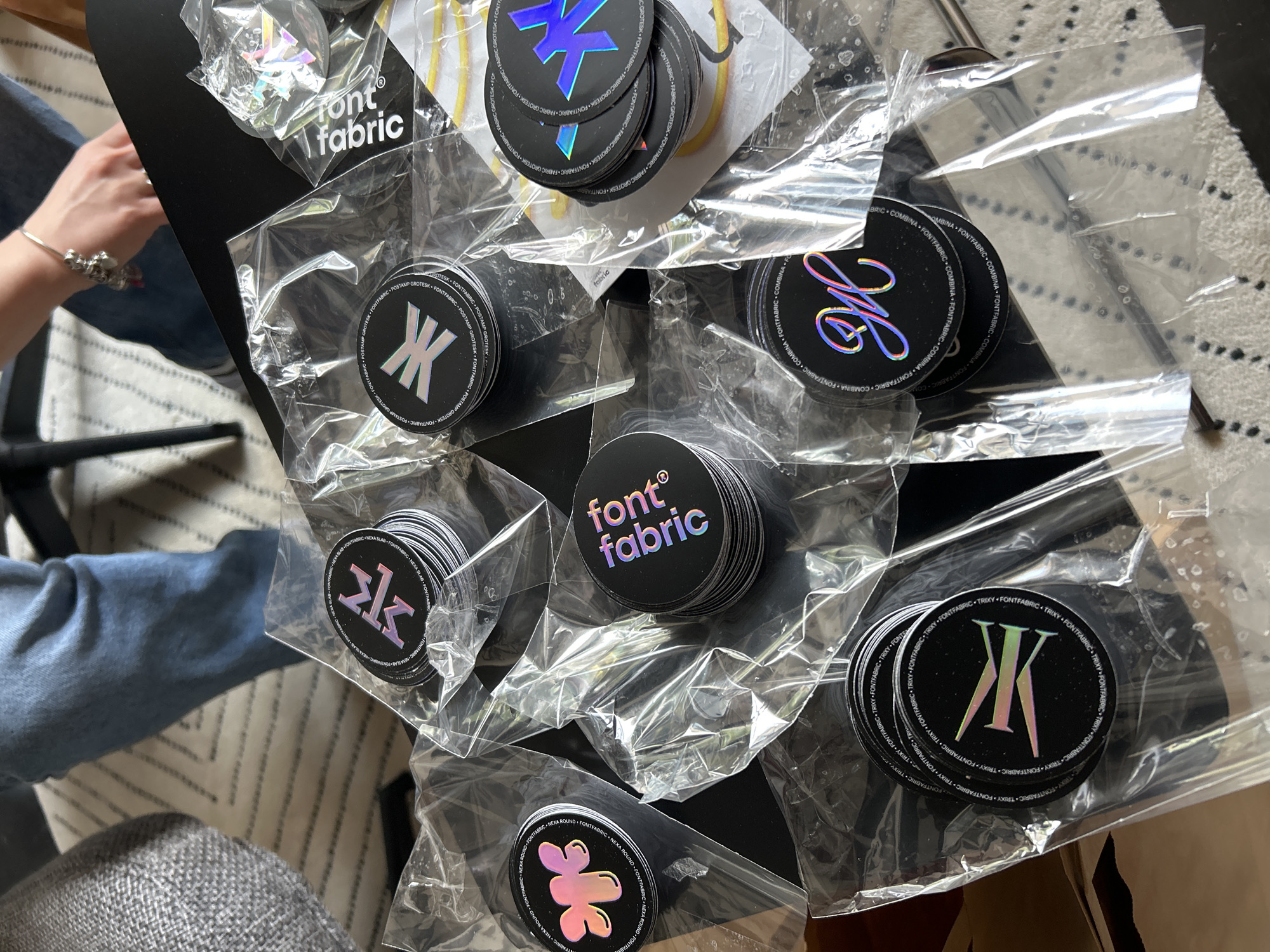 A high-angle shot displays a collection of round, black stickers, each stacked in small piles and individually wrapped in clear plastic bags. The stickers are scattered on a black table and each features a unique, vibrant, holographic design. Around the edge of the stickers are printed with the texts of different font names such as “POSTAMP GROTESK”, “GROTESK”, “NEXA SLAB”, "TRIXY", "ROUND" and “COMBINA”. On some stickers feature the words "font fabric" in a distinctive typeface. A person wearing a bracelet and jeans is visible in the top left corner of the frame. The environment is partially obscured by the objects in focus, revealing a section of a patterned rug. The location is indeterminable, but the style of the stickers suggests they might be related to a business with a font design/typography focus. Bulgaria is indeterminable.