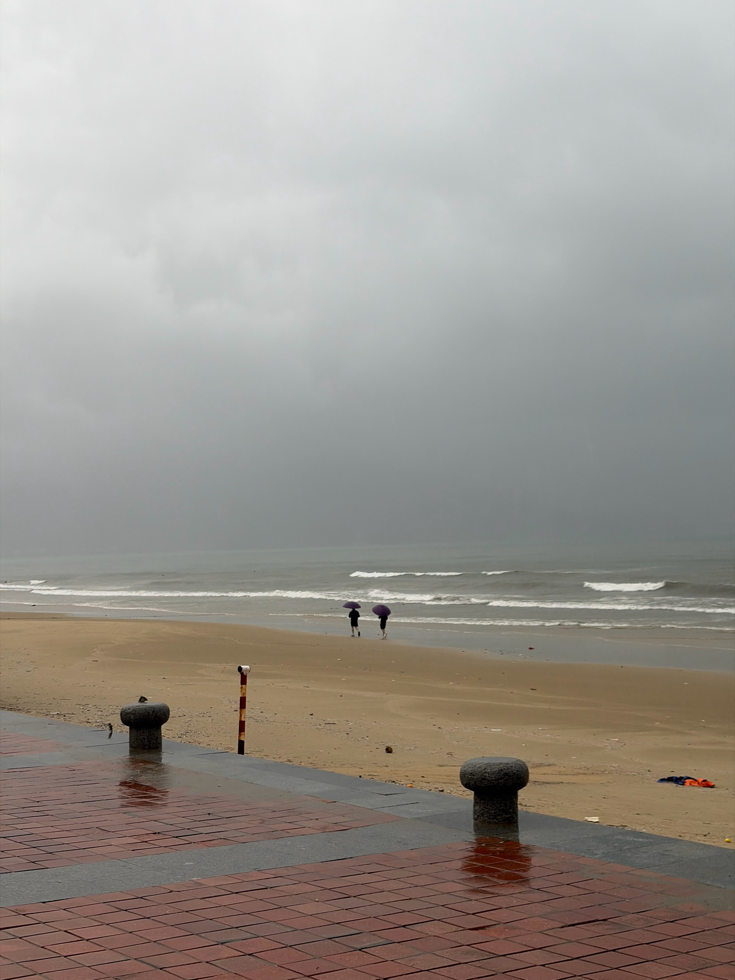 This is a photograph of the My Khe beach in Da Nang, Vietnam, on a cloudy and possibly rainy day. The sky is a uniform gray, filled with heavy clouds. The ocean is a muted gray-blue, with moderate waves breaking on the shore, leaving white foam on the wet sand. The beach is mostly empty, with a few seagulls visible on the sand. In the distance, two people are walking on the beach, both holding dark umbrellas. In the foreground, a paved walkway made of red bricks and gray stones is visible. Several bollards are placed along the edge of the walkway, one of which has a bird perched on top. A striped pole, red and white, stands near the bollard. The walkway is wet, reflecting the overcast sky and surrounding elements, indicating recent rainfall. Some debris, including what appears to be a blue and orange piece of clothing or tarp, is scattered on the sand near the edge of the walkway.