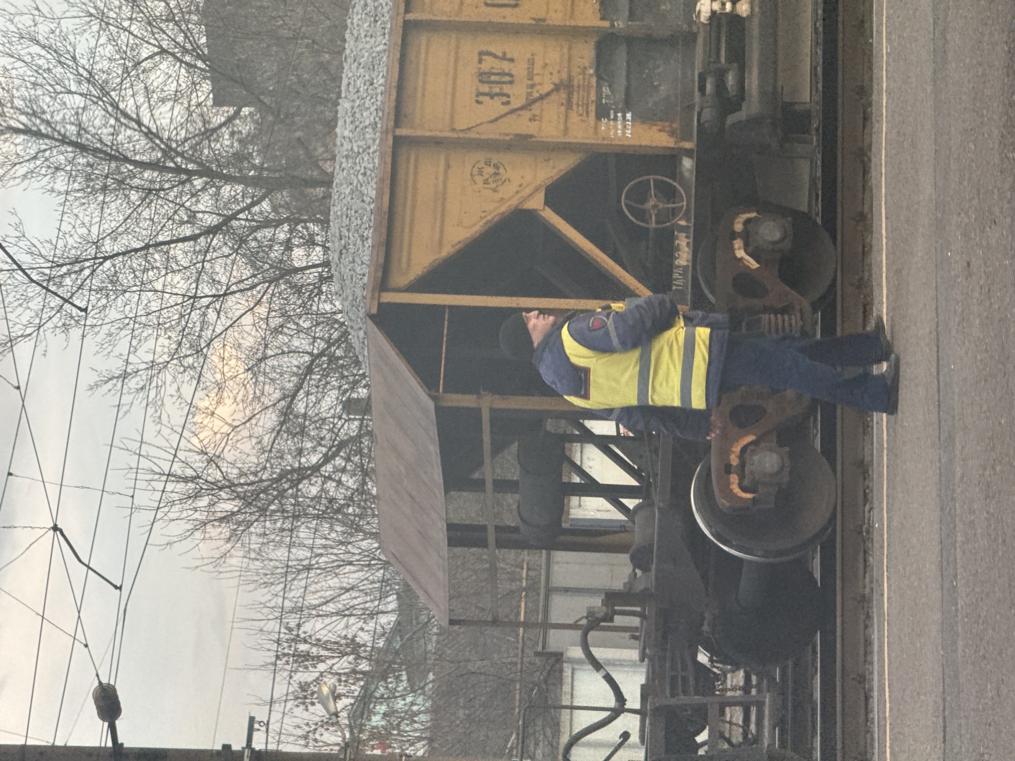 An outdoor scene depicts a rail worker inspecting a railcar, likely in or near Gryazi, Russia. The railcar is a yellow hopper car loaded with what appears to be gravel or ballast. The worker is wearing a dark jacket under a yellow safety vest and dark pants. He is standing next to the railcar's wheels, looking upwards towards the car's body. The environment includes overhead power lines, bare trees indicating winter or early spring, and what may be a building in the background. The lighting suggests an overcast day.