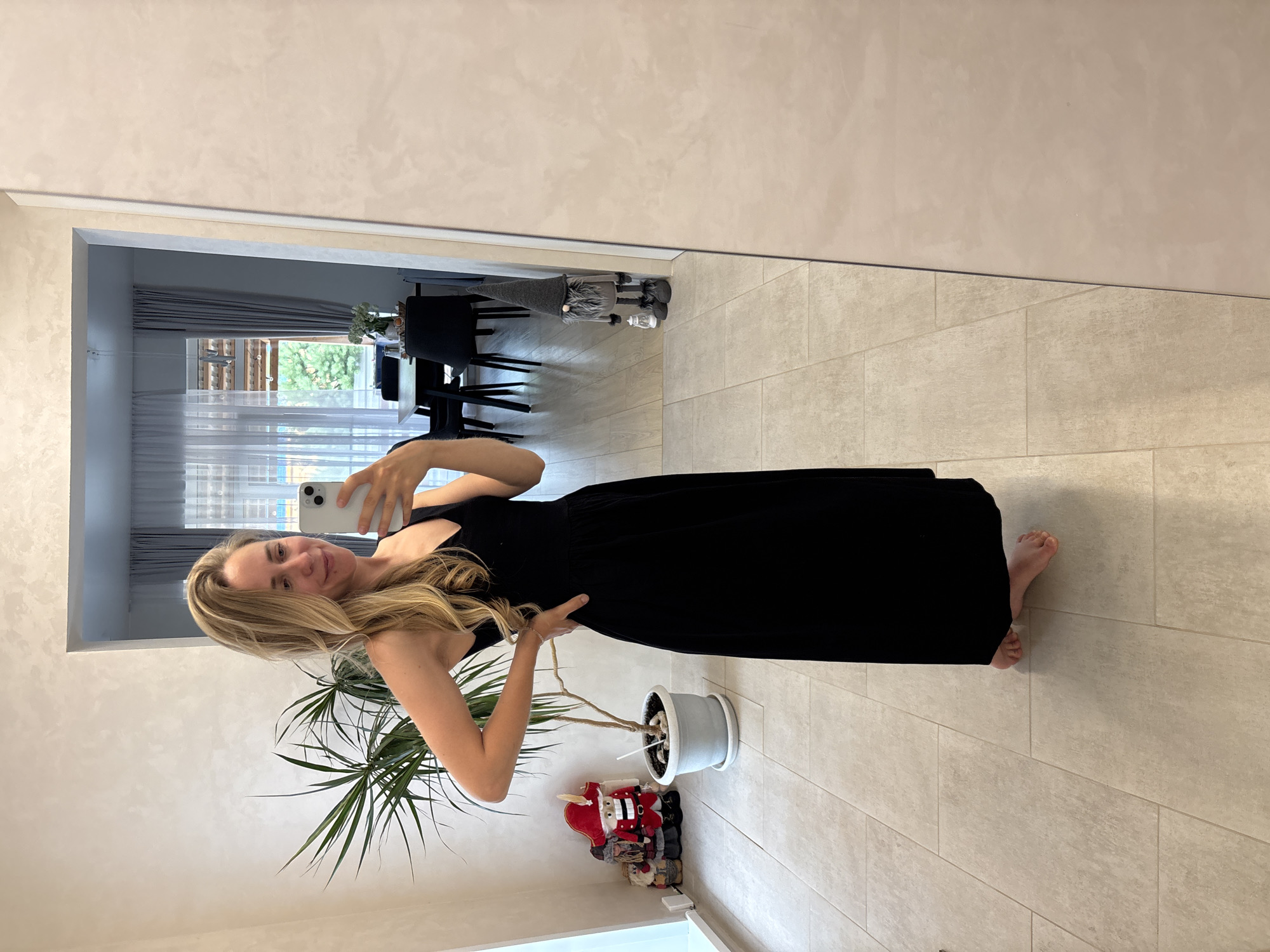 A blonde woman wearing a black sleeveless maxi dress stands in an indoor space, taking a mirror selfie with her phone. She is barefoot, with her left hand on her hip. To her left, a tall potted plant with thin, spiky leaves is visible, next to two decorative gnome-like figures dressed in red and blue. In the background, through an open doorway, a dining area with a table and chairs can be seen, along with curtains and a window offering a view of trees and the sky. The floor is tiled. The scene appears to be an interior of a home. The location of Chaikovsky, Russia is not discernible from the visual information provided in the image.