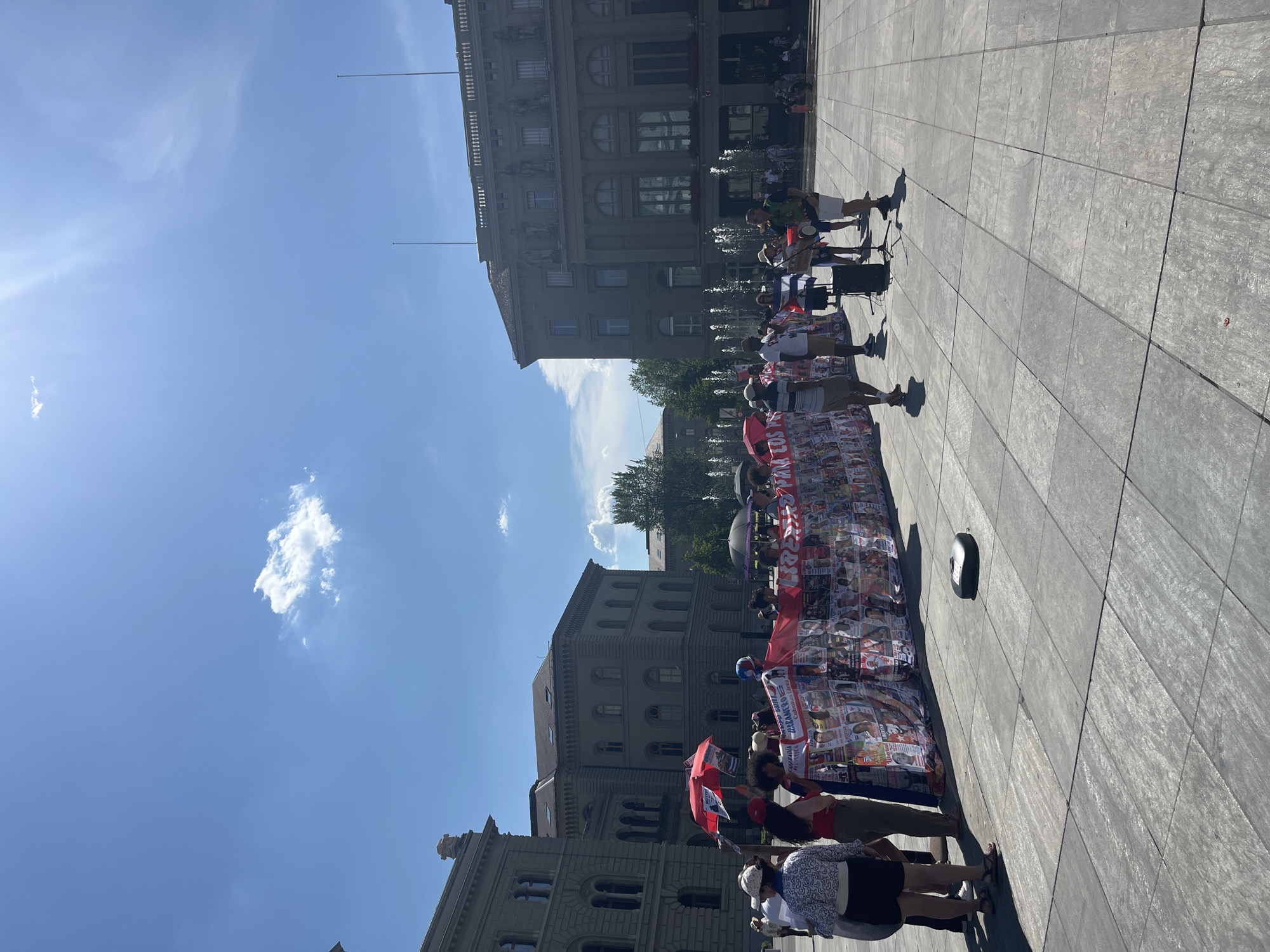 The image depicts a daytime outdoor scene in the city of Bern, Switzerland, likely in front of the Federal Palace (Bundeshaus). Several people are gathered in a paved plaza. In the foreground, a group of individuals stands behind a long banner adorned with numerous photographs and text, some of which is in Spanish. To the right of the banner, a small group is gathered around a man playing a drum. In the background, large, ornate buildings with classical architecture are visible, along with fountains and trees. The sky is blue with scattered clouds and bright sunlight. The overall atmosphere appears to be that of a public gathering or demonstration.
