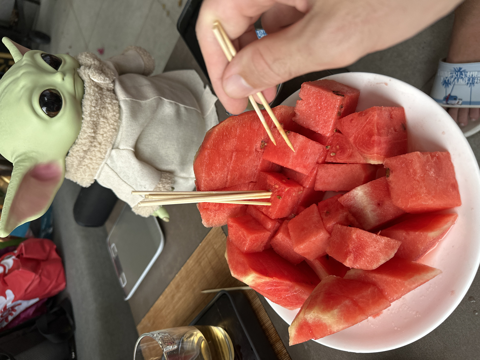 A plate of cut watermelon is on a table, with a hand holding toothpicks to pick up a piece of watermelon. A toy figurine of Grogu from Star Wars is in the background.