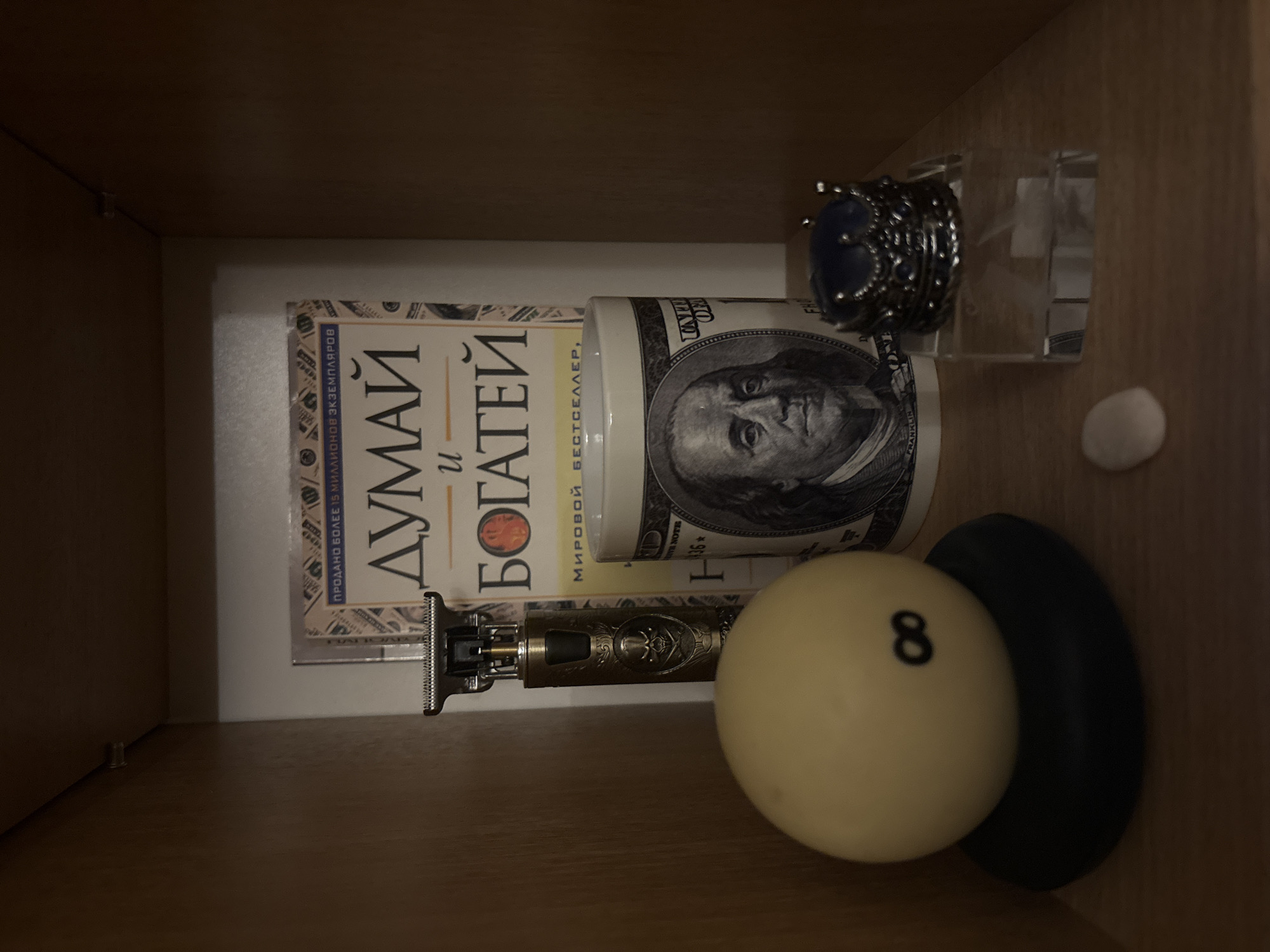 A collection of items is displayed on a wooden shelf. From left to right, there is a pool ball with the number "8" on it, resting on a dark, circular base. Next to it is a metallic electric trimmer with a skull design. Behind the trimmer and pool ball, a book with a golden cover and Cyrillic text is partially visible. To the right of the book is a white mug featuring the portrait of Benjamin Franklin and dollar bill designs. In front of the mug, a small, transparent perfume bottle with a decorative crown-shaped cap is placed. To the right of the pool ball and slightly in front of the mug, a small, light-colored stone is visible on the shelf. The background appears to be the interior of a wooden cabinet or shelf. The lighting is dim, casting shadows on the objects.