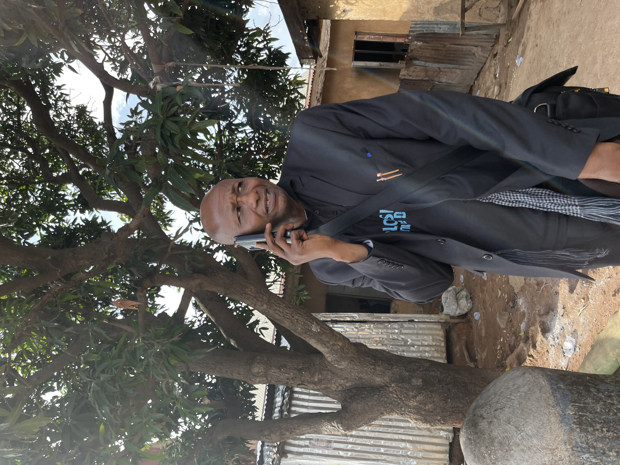 The image depicts a man in a dark jacket speaking on a mobile phone outdoors. The location is indicated as Jos, Nigeria. He stands near a tree with dense foliage, and behind him is a building with a visible window. The ground appears to be dirt with some debris. The man has a bald head and is wearing a black blazer and a scarf with a checkered pattern is visible. A black bag is visible behind the man. A rainbow glint is visible above his jacket.