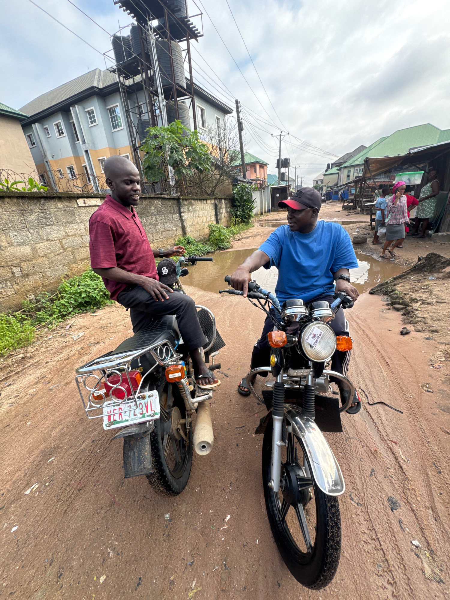 Two men are on motorcycles in Ihiagwa, Nigeria. The man on the left is wearing a maroon polo shirt and dark pants, sitting on a motorcycle with a license plate that reads "HER 729YL". The man on the right is wearing a blue t-shirt and a red and black baseball cap, sitting on a motorcycle with a circular sticker on the headlight that appears to be a seal. Both men are looking towards each other and appear to be engaged in conversation. The ground is muddy and appears to be a dirt road, with puddles of water. In the background, there are buildings, some of which are multi-story, and a market scene with people and stalls. The sky is overcast.