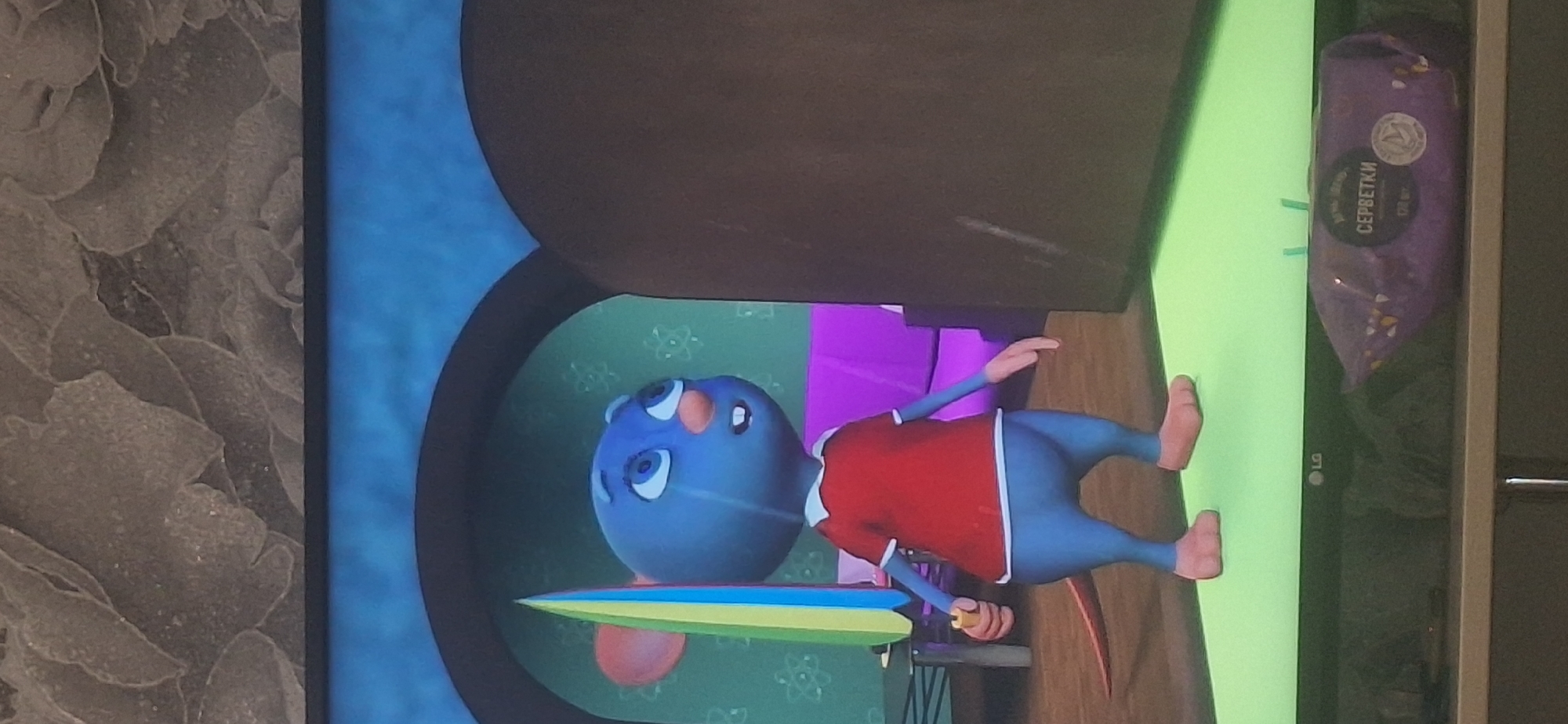 A blue cartoon mouse character, wearing a red shirt with a white collar and cuffs, and blue pants, stands in what appears to be a living room. The mouse is holding a colorful, striped object, possibly an umbrella or a prop. In the background, a purple sofa is visible, and the walls have a floral pattern. To the right of the mouse is a large, dark brown, rounded object, possibly a doorway or a piece of furniture. The scene is captured on a television screen, as indicated by the LG logo visible on the bottom bezel. A package of wet wipes with Cyrillic lettering is placed on the shelf below the television. The image does not contain any information that would allow for its location to be determined in Bila Tserkva, Ukraine.