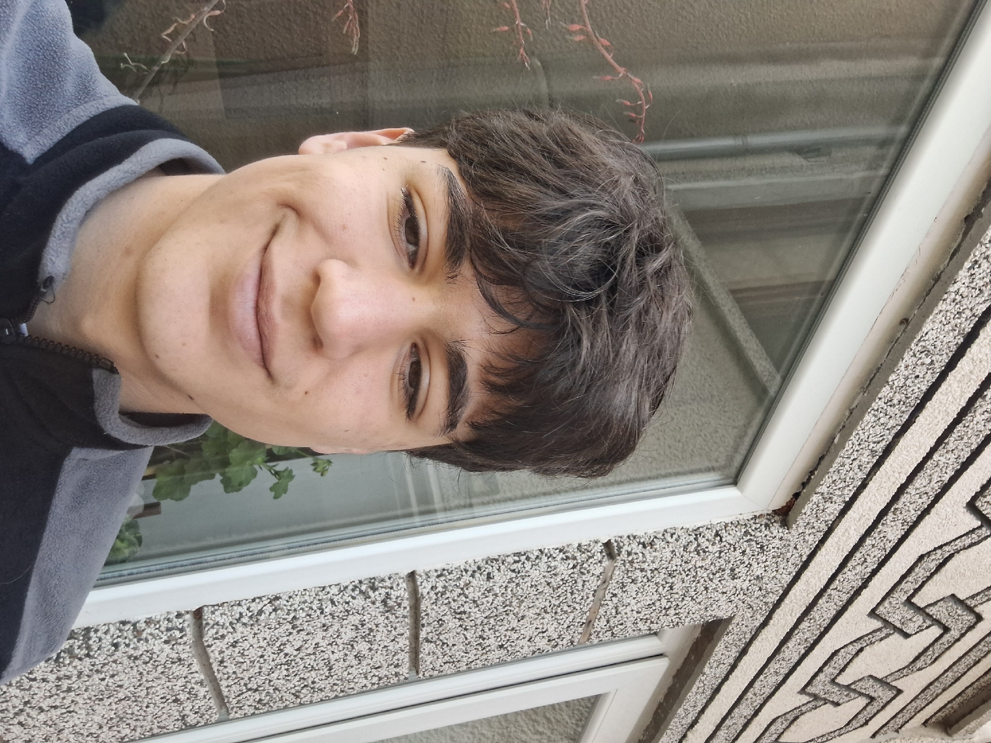 A selfie photograph shows a person with dark hair, wearing a black and grey zippered jacket. The person is smiling slightly. The background appears to be a window, with what may be plants visible through the glass. Outside the window, a textured, possibly stone or stucco, surface is visible. The pattern on the outside surface has a maze-like design. There is no indication of the location being Stara Zagora, Bulgaria from the given information.