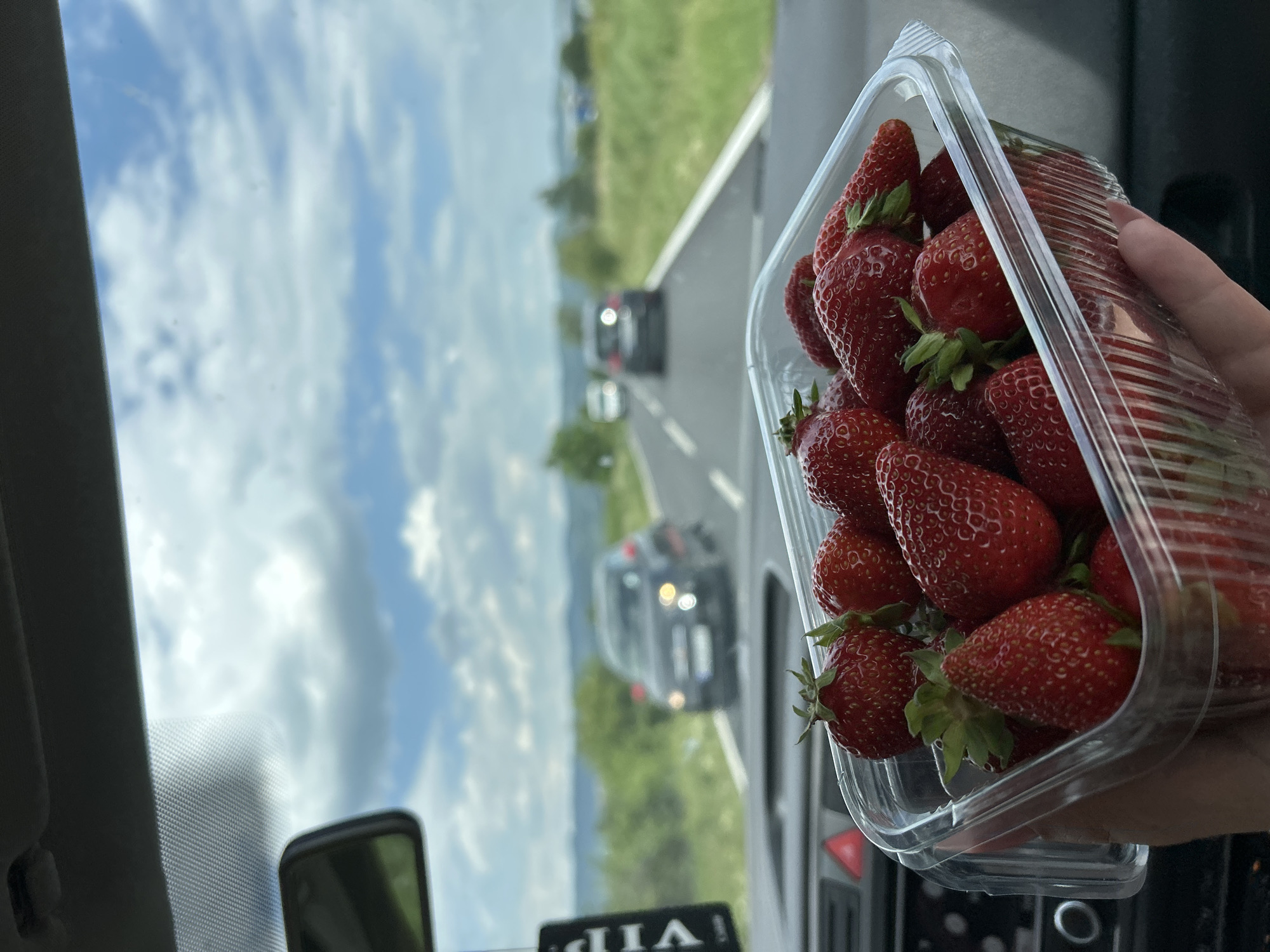 A close-up shot taken from inside a vehicle shows a clear plastic container filled with fresh, ripe strawberries. The container is held in someone's hand, positioned in the foreground of the image. Through the vehicle's window, a blurry landscape is visible, featuring a road with moving cars, green fields, and a sky with scattered clouds. The location appears to be Dolna Banya, Bulgaria, although this is based on contextual information and not directly discernible from the image alone.