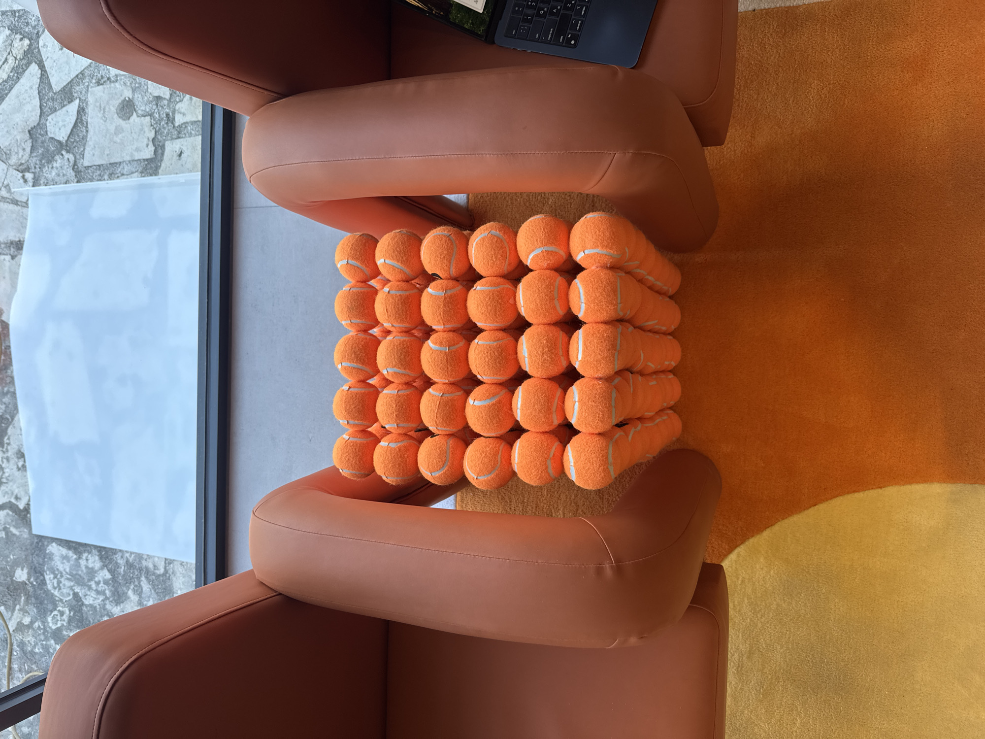 An overhead shot captures a pile of bright orange tennis balls neatly stacked between two plush, burnt-orange armchairs. The tennis balls are arranged in a roughly rectangular prism formation, with layers of five balls each. Beyond the armchairs, a large window overlooks a paved outdoor area and a bright blue sky. To the right of the frame, a laptop rests on the arm of one of the chairs, its screen displaying an image. The scene is set on an orange and yellow rug, suggesting an indoor space. The location is a living room or lounge area in Kuta Selatan, Indonesia.