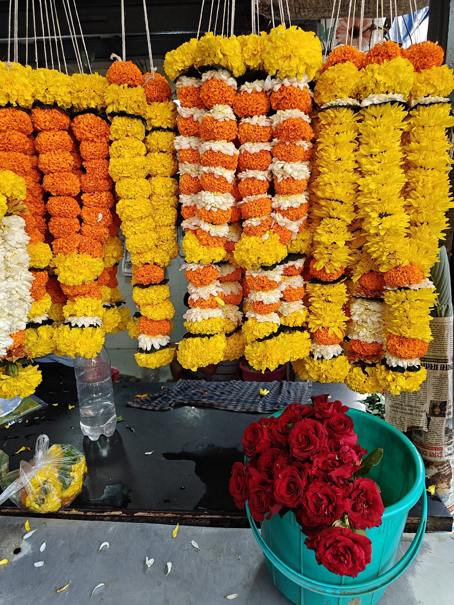 The image depicts a flower stall, likely in Mumbai, India, based on the language visible on the newspaper. Numerous flower garlands dominate the scene, hanging in rows. They are made of combinations of yellow, orange, and white flowers, often alternating colors. Below the garlands, on what appears to be a countertop, is a green plastic bucket filled with red roses. A plastic bag with yellow flowers is also visible. Debris, including fallen petals, litters the surface. Other items are also on the countertop, including a water bottle.