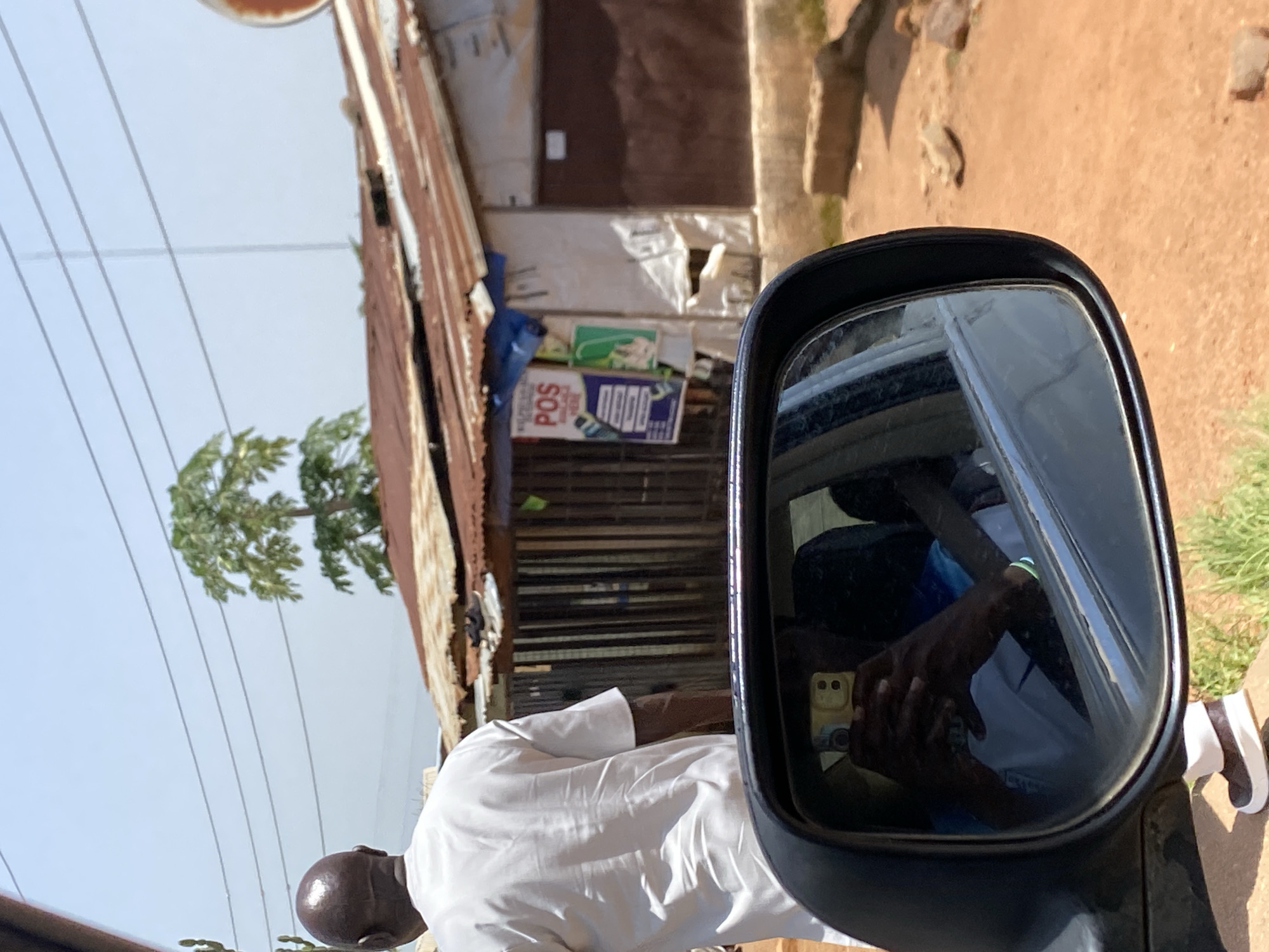 The image depicts a scene in Gura Top, Nigeria, focusing on a man walking away from the camera, dressed in a white kaftan. His back is to the viewer, and his head is bald. In the foreground, a car's side mirror reflects the interior of the car. Inside the mirror, a person is visible, holding a yellow smartphone and appearing to take a selfie. The person is wearing a blue and white shirt. Outside the car, a simple building with a corrugated iron roof and a wooden fence is visible. A tree with large leaves stands behind the building. The ground is covered in reddish-brown dirt and some sparse green vegetation. Utility wires are strung across the sky. A sign with "POS" is posted on the building.