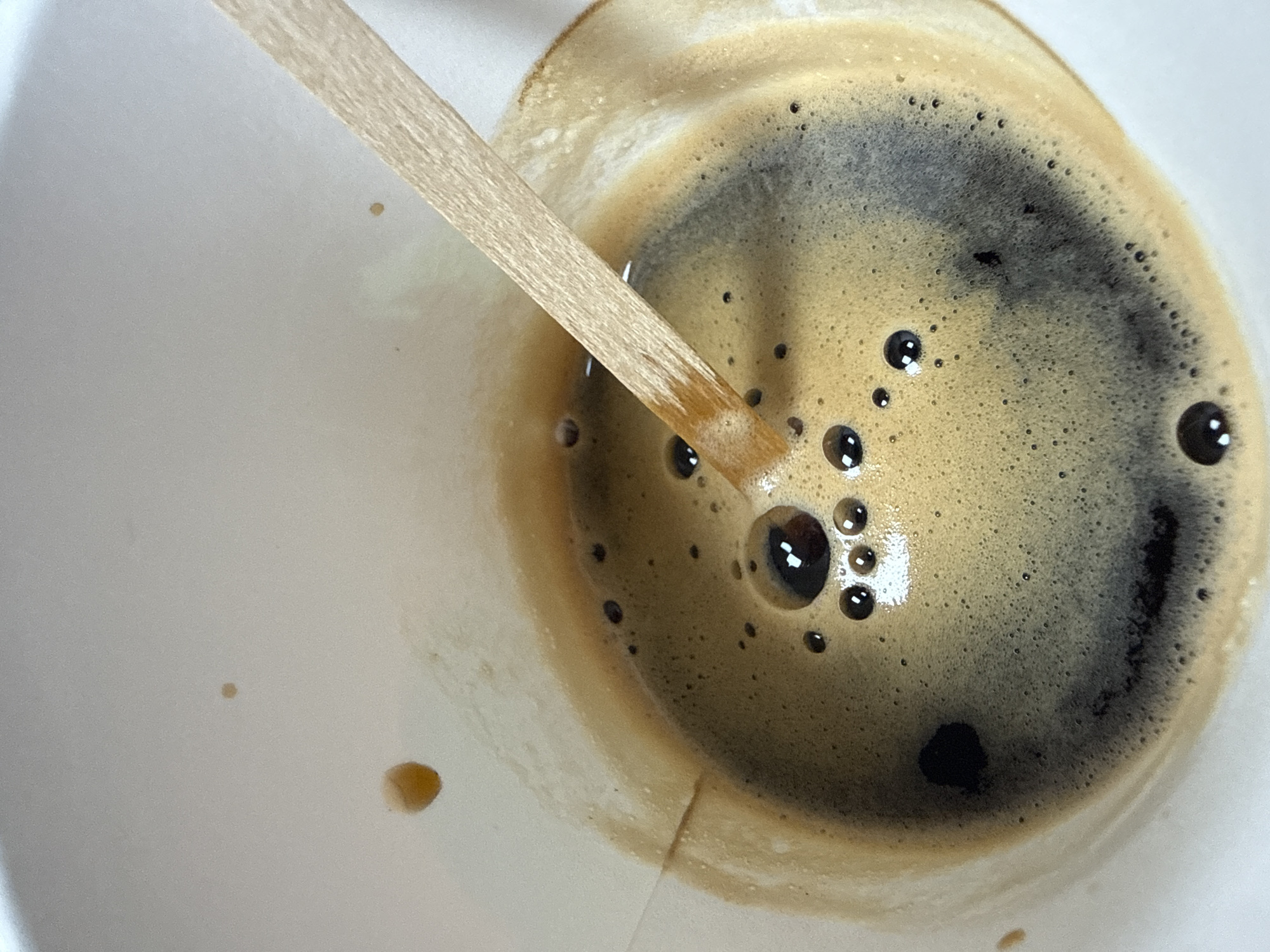 A close-up overhead shot shows a wooden stirring stick partially submerged in a cup of coffee. The coffee has a thick layer of crema, with small bubbles scattered across its surface. There are also a few dark droplets of coffee on the white rim of the cup, suggesting some spillage. The background is out of focus and appears to be a plain white surface. The scene is likely indoors, possibly in a cafe or home setting. There is no indication of the city of Aksakovo, Bulgaria in the image.