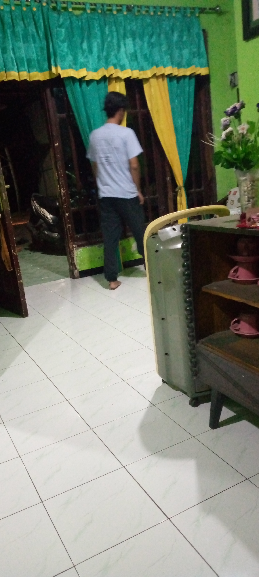 A person, viewed from behind and in motion, is standing in what appears to be a residential interior in Sumberwono, Indonesia. The individual is wearing a light-colored t-shirt with a graphic design on the back and dark pants. They are barefoot and are facing a doorway with curtains partially drawn. The curtains are a vibrant combination of emerald green and yellow, suggesting a decorative element within the room.
The environment features tiled flooring, which is light-colored with a subtle pattern. To the right of the frame, a piece of furniture, possibly a cabinet or shelf unit, is visible. It has a dark wooden finish with shelves containing a few pink items that resemble cups or bowls. Next to this furniture is a large, light-green object with a textured, ribbed side, possibly an appliance or part of a larger structure.
The background beyond the doorway shows a dark interior space, with a glimpse of what could be a motorcycle on the left. The walls are painted green, and the lighting suggests it might be evening or a dimly lit room. The overall scene captures a candid moment within a domestic setting.