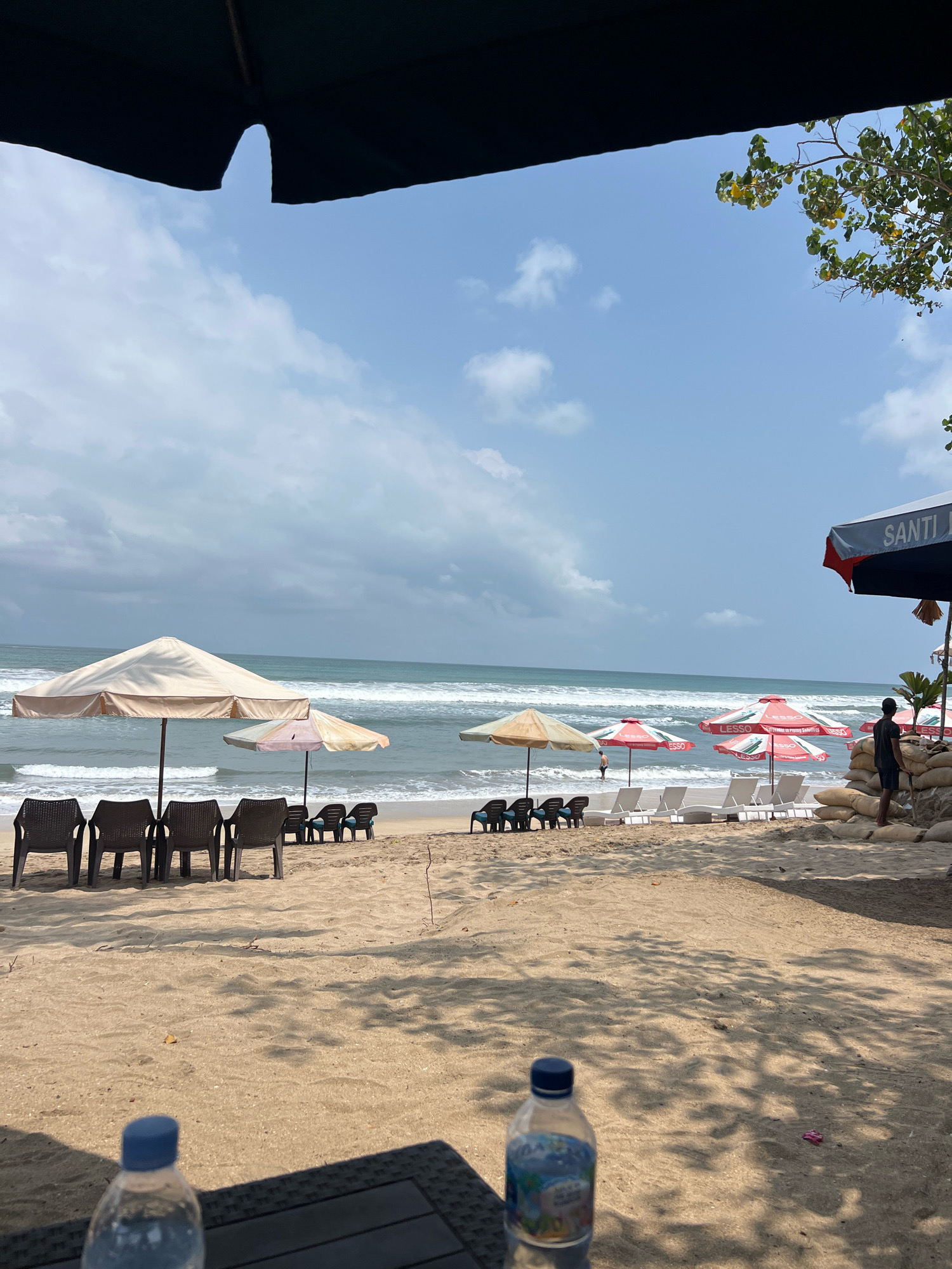 The image depicts a beach scene in Kuta, Indonesia, under a partly cloudy sky. In the foreground, a black table with a bottle of water is visible, casting a shadow. The middle ground features a sandy beach lined with beach chairs and umbrellas. Several people are present on the beach, some relaxing on loungers and others wading in the ocean. In the background, the sea stretches out towards the horizon. The overall atmosphere is serene and idyllic, with the gentle waves of the ocean and the warm sunlight contributing to the peaceful ambiance. The location is identified as Kuta, a popular tourist destination in Indonesia known for its beautiful beaches and vibrant atmosphere.