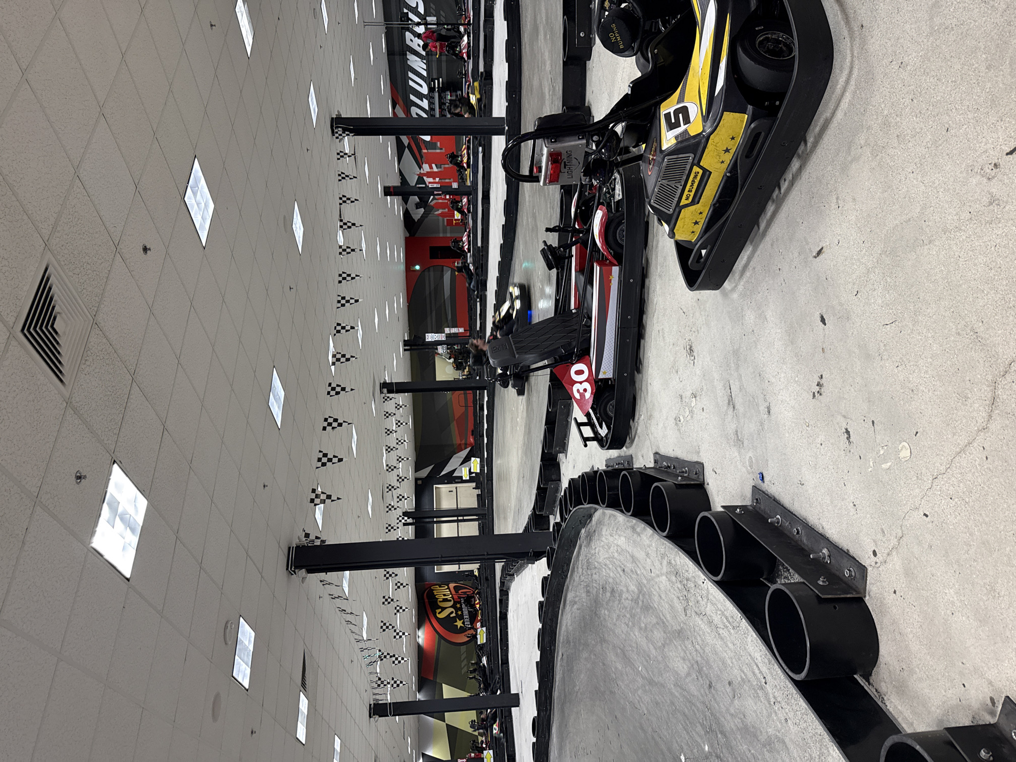 The image shows an indoor go-kart racing track. Several go-karts are visible, some with drivers in them, staged on the track. The track is lined with black barriers. Checkered flags decorate the track's surroundings. A sign with the words "Scene 75 Entertainment" is partially visible. The general appearance and branding suggest the location is Scene75 Entertainment Center in Columbus, Ohio. The lighting is bright and appears to be from overhead fluorescent fixtures on the ceiling. The floor is concrete.