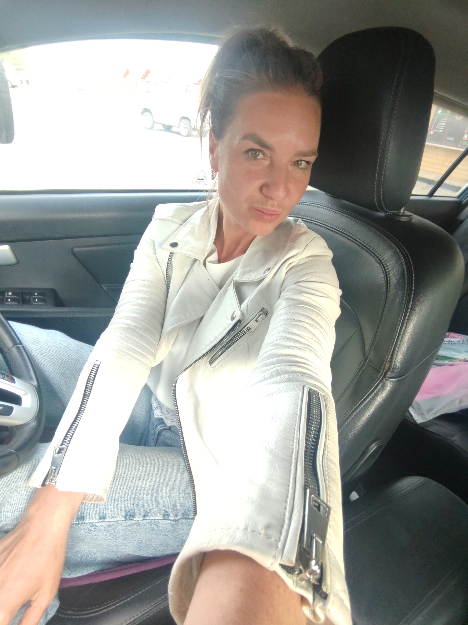 A woman is sitting in the driver's seat of a car, looking at the camera with her lips pursed. She is wearing a white leather jacket over a white top and blue jeans. The interior of the car is visible, including the steering wheel, dashboard, and leather seats. The scene appears to be in Petropavlovsk-Kamchatsky, Russia, based on visual cues in the background which suggest a Russian urban or suburban environment with typical Russian automobiles. The lighting suggests it is daytime, possibly overcast.