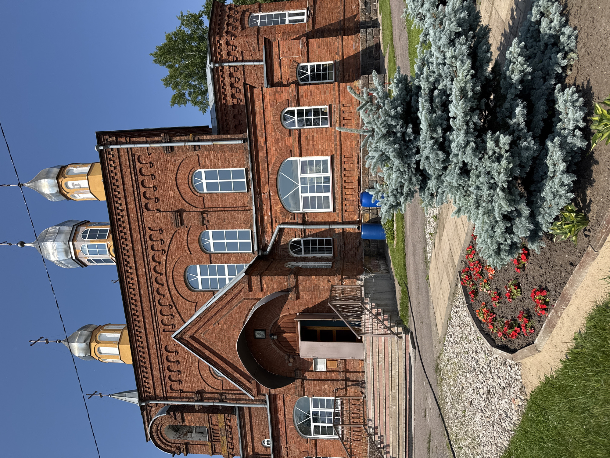 The image depicts the exterior of an orthodox church constructed from red brick in the city of Chudovo, Russia. The church features multiple domes topped with crosses, arched windows, and an entrance with a covered portico. In the foreground, a landscaped area with a small blue spruce tree and red flowers is visible. The sky is clear and blue. There are no people or animals present in the image.