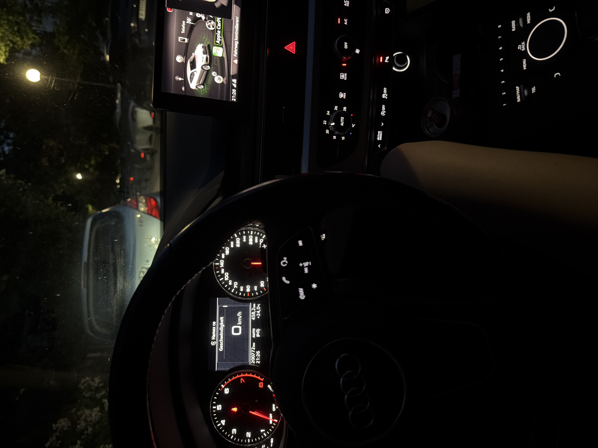 An interior view of an Audi car is depicted at night, taken from the driver's perspective in Ruse, Bulgaria. The car is stationary, with the instrument cluster displaying 0 km/h, an odometer reading of 200772 km, the time as 21:26, and an outside temperature of +24.0°C. The dashboard language is set to German, showing "Geschwindigkeit" (speed) and "READY" illuminated. A red warning indicator is active on the left side of the cluster.
The central MMI screen also shows the time 21:26, features Apple CarPlay, a graphical representation of a white Audi coupe, and a warning message "Fahrweg kontrollieren!" (Check driving path!). Below the screen, the climate control panel indicates "AUTO" mode with a temperature setting of 22°C. A hazard light button (red triangle) is visible. Further down the center console, a Red Bull can is situated in a cup holder, next to illuminated controls for "drive select," parking brake, and the MMI rotary controller with associated "NAV/MAP," "TEL," "MENU," "BACK," "RADIO," and "MEDIA" buttons. A light-skinned arm or leg is partially visible on the right side of the frame.
Through the windshield, a dark urban environment at night is visible. Several vehicles are parked or dimly seen in the distance, including the rear of a light-colored car directly in front of the Audi. Two spherical streetlights illuminate the scene, casting light on trees and dark vegetation. The windshield exhibits minor streaks, suggesting recent moisture or condensation.