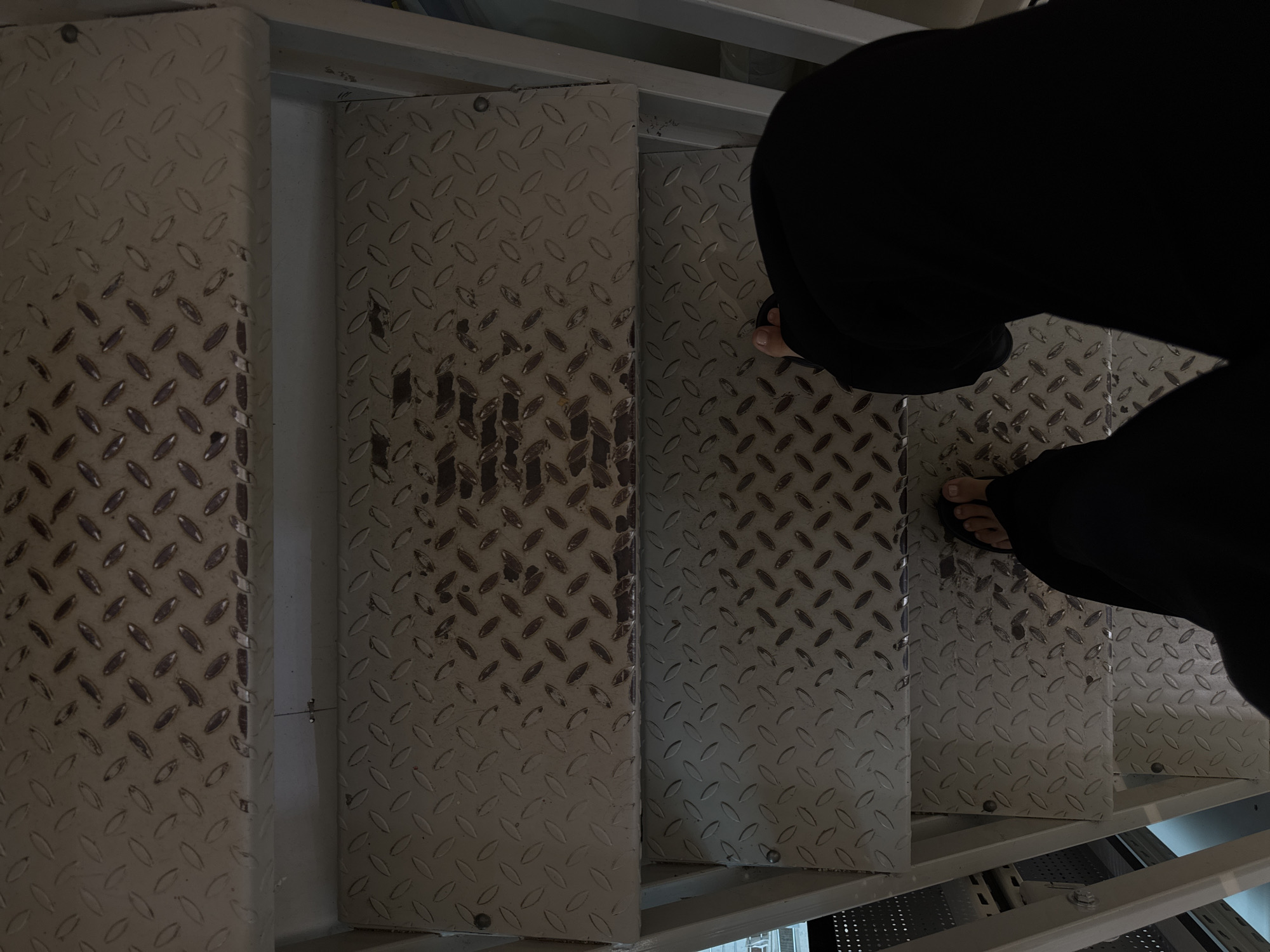 A first-person perspective shot looking down at a set of metal diamond plate stairs. A person is wearing black pants and dark sandals, with their feet visible on the lower steps. The stairs are worn and appear to be in an industrial or utilitarian setting. There are no identifiable Moscow landmarks.