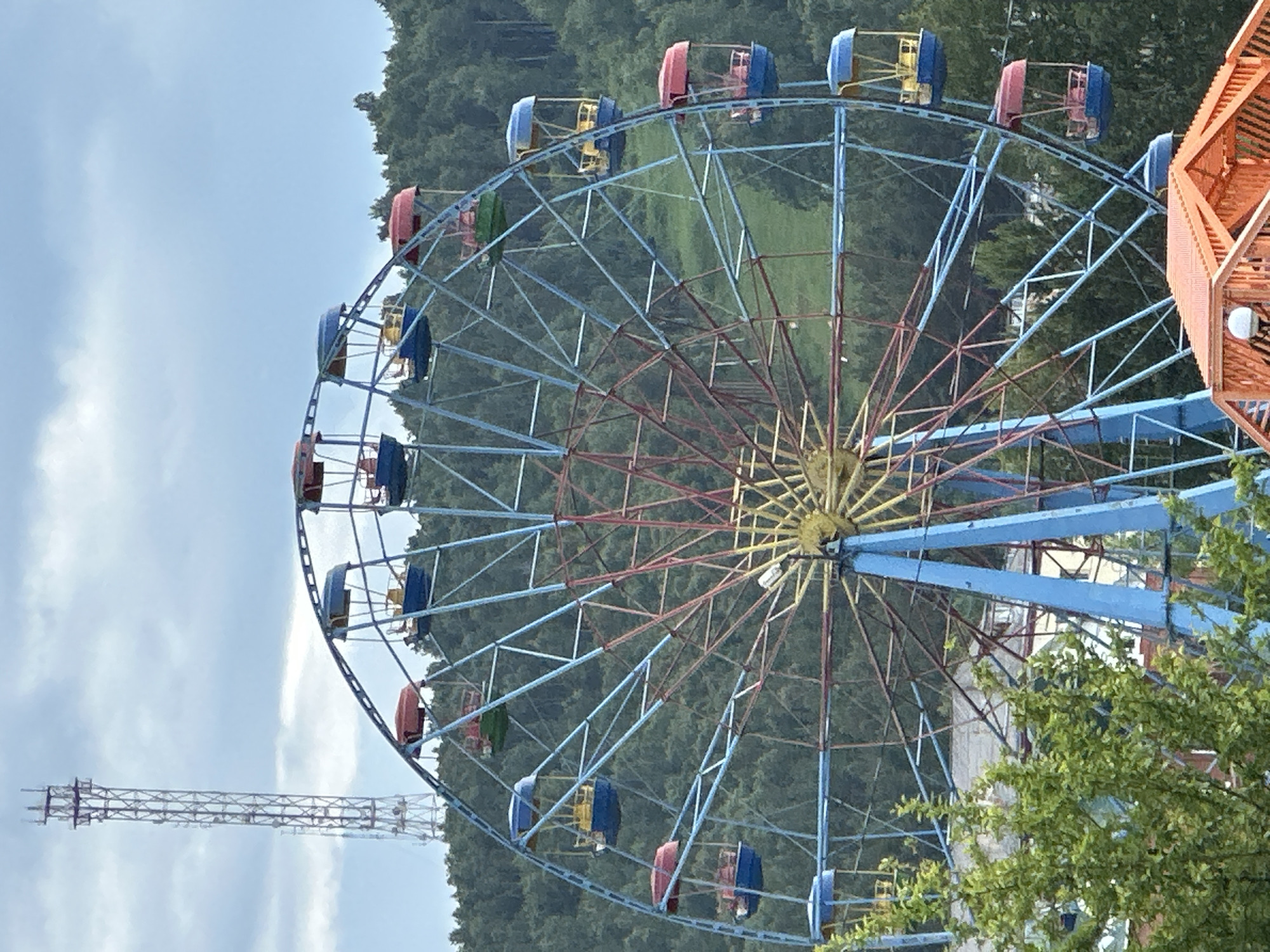 A ferris wheel with blue and red gondolas is situated in front of a densely wooded hillside under a cloudy sky. A tall, lattice-like telecommunications tower is visible in the distance to the left of the ferris wheel. The ferris wheel has a central yellow hub and red and blue spokes. Some of the gondolas are red-topped, others are blue-topped, and at least one is green-topped. The lower portion of the ferris wheel is supported by blue legs. In the foreground, a portion of an orange structure, possibly a bridge or walkway, is visible. The overall scene appears to be in a park or amusement area.