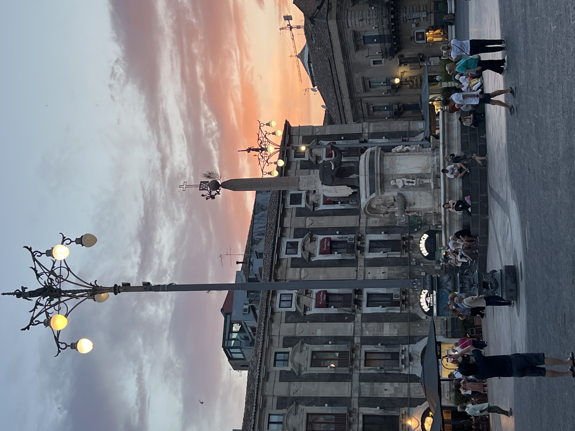 The image shows a street scene in Catania, Italy, specifically Piazza Duomo, at dusk. A street lamp with multiple globes stands prominently in the foreground, casting a warm light. Behind it, the Fontana dell'Elefante, a central monument featuring an elephant sculpture, is visible. The sky is filled with dramatic clouds tinged with hues of orange and pink from the setting sun.
Several people are present in the plaza. In the mid-ground, a man is taking a photo with his phone. Other individuals are seen walking, sitting on the steps of the fountain, or standing in small groups. Some buildings with traditional architecture line the square, featuring balconies and arched windows. The overall atmosphere suggests a lively public space in the early evening.