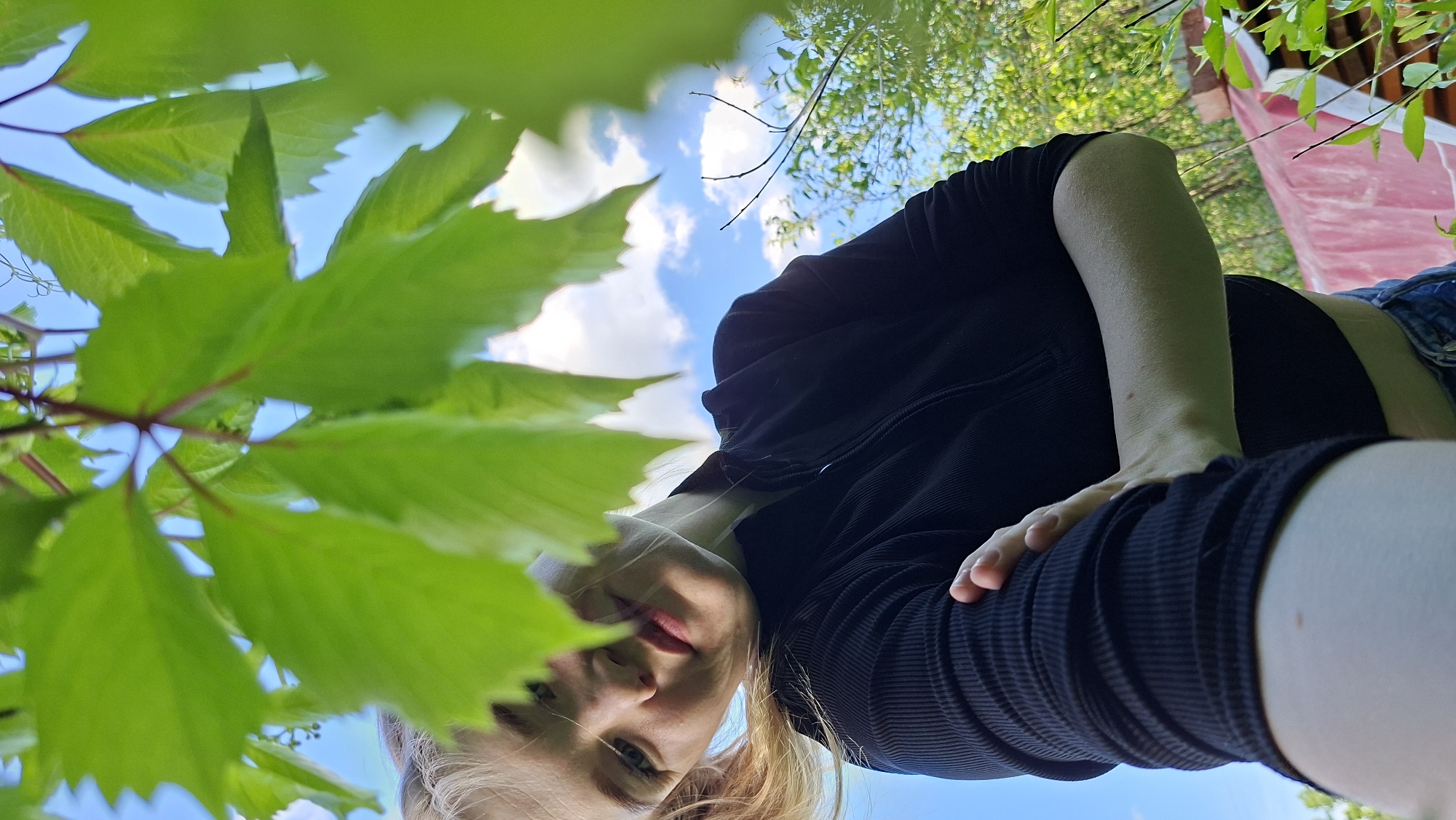The image is a low-angle shot taken outdoors. A person, presumed to be a young woman, is looking down at the camera. She has fair skin, light-colored hair, and is wearing a dark, ribbed top and denim shorts. Her hand is resting on her thigh. The background consists of green foliage and a blue sky with scattered clouds, suggesting a bright, sunny day. A glimpse of a red structure is visible in the background, possibly part of a building. The location is described as being in the city of Большой Букор, Russia. The angle of the photo suggests it was taken from ground level, looking upwards.