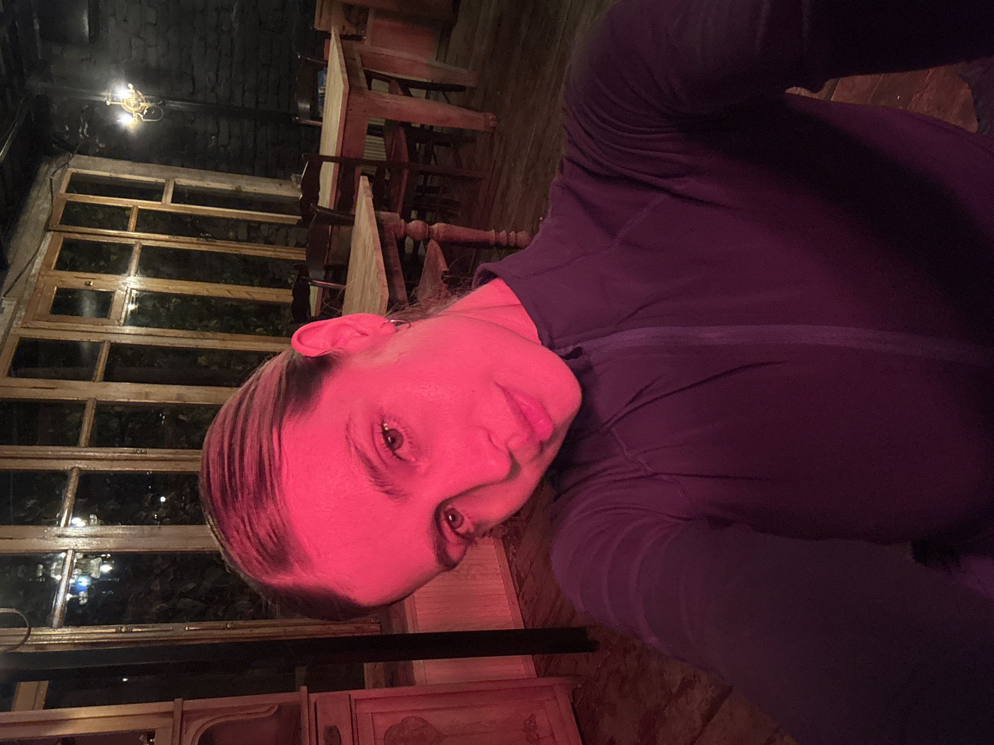 A close-up selfie, captured from a tilted perspective, features a young adult, likely female, whose face is bathed in a vivid red light. She is looking directly at the camera with a subtle expression, her light-colored eyes prominent despite the strong red hue. She is wearing a dark purple long-sleeved top with a zipper on the front.
The background suggests an intimate, dimly lit interior, possibly a cafe or restaurant in Tbilisi, Georgia. To the viewer's right, several wooden tables and chairs are visible, some appearing stacked. Behind the person is a large, multi-paned wooden window, through which dark foliage and faint background lights are discernible, indicating it is likely evening or night. A decorative, brass-colored chandelier or light fixture hangs from the dark ceiling above the window. In the lower left corner, a portion of a wooden cabinet with ornate details can be seen. The overall impression is one of a moody and somewhat dramatic setting, emphasized by the contrasting red light on the subject against the warm, subdued tones of the wooden decor. No visible text is present in the image.