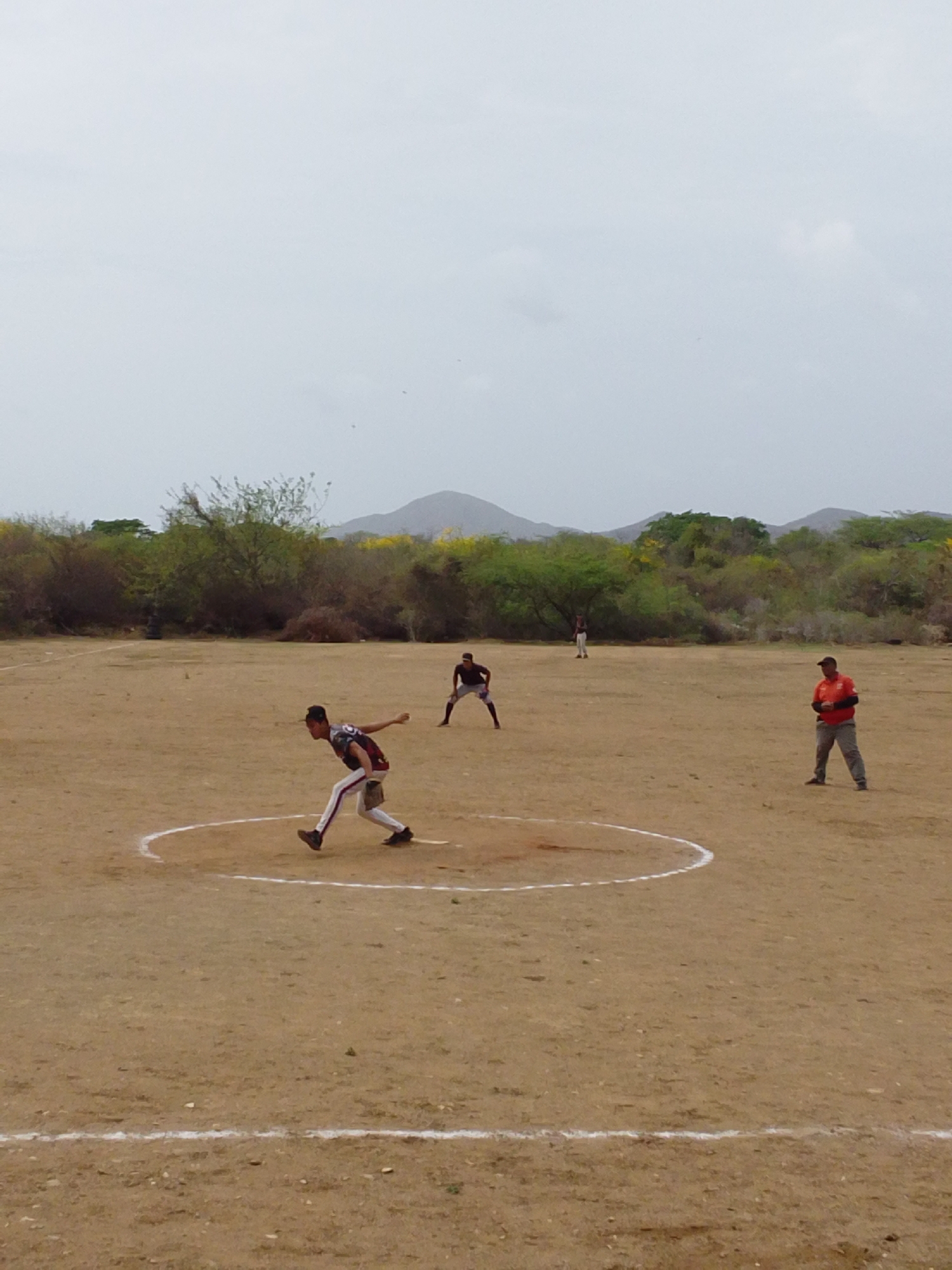 The image captures an outdoor baseball or softball game/practice on a dry, light brown dirt field. A youthful male player, wearing a dark jersey with white and red accents, white pants, and dark shoes, is positioned within a white chalk circle in the left-center foreground, captured mid-pitch with his right arm extended backward. His left hand, wearing a glove, is partially visible. To the left and slightly behind the pitcher, another male individual in a dark shirt and pants is crouched in a ready stance. Further back and slightly to the right, a third male player in a light shirt and dark pants stands upright. On the far right, a fourth male individual, wearing an orange long-sleeved shirt, dark cap, and light pants, observes the scene with his hands clasped in front of him. A straight white chalk line extends horizontally across the lower portion of the frame, indicating a field boundary. The background consists of a dense band of green and brown trees and bushes, with some displaying bright yellow blossoms, giving way to hazy, blue-gray mountains under a uniformly light gray, overcast sky. No specific city or state can be determined from the visual information.