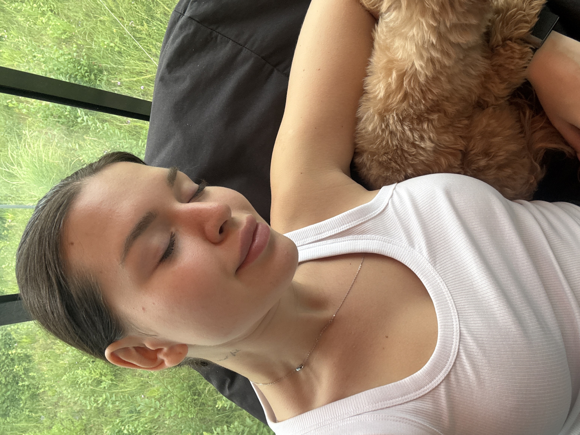 In a candid, close-up shot taken from a slightly high angle, a young woman with dark hair pulled back rests her eyes, her head tilted slightly downwards. She wears a white ribbed tank top and a delicate silver necklace. Her right arm is bent, and she appears to be holding or leaning against a small, fluffy, light brown dog. The background is out of focus, showing lush green foliage and a dark vertical element, possibly a window frame or architectural detail. The setting appears to be indoors with a view of an outdoor natural landscape. This image was not taken in Prague, Czechia.