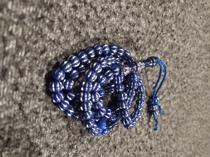 A single blue and silver-striped tasbih (prayer beads) is resting on a dark grey, textured surface, likely a carpet or fabric. The tasbih consists of numerous spherical royal blue beads, each featuring multiple horizontal silver-colored stripes. A prominent blue cylindrical imam bead, adorned with silver-colored decorative bands, is visible at the apex of the strand, from which a short, blue braided cord tassel extends. The string of beads is coiled loosely on the surface. No individuals or other objects are present in the scene. The location is Unknown, Unknown.