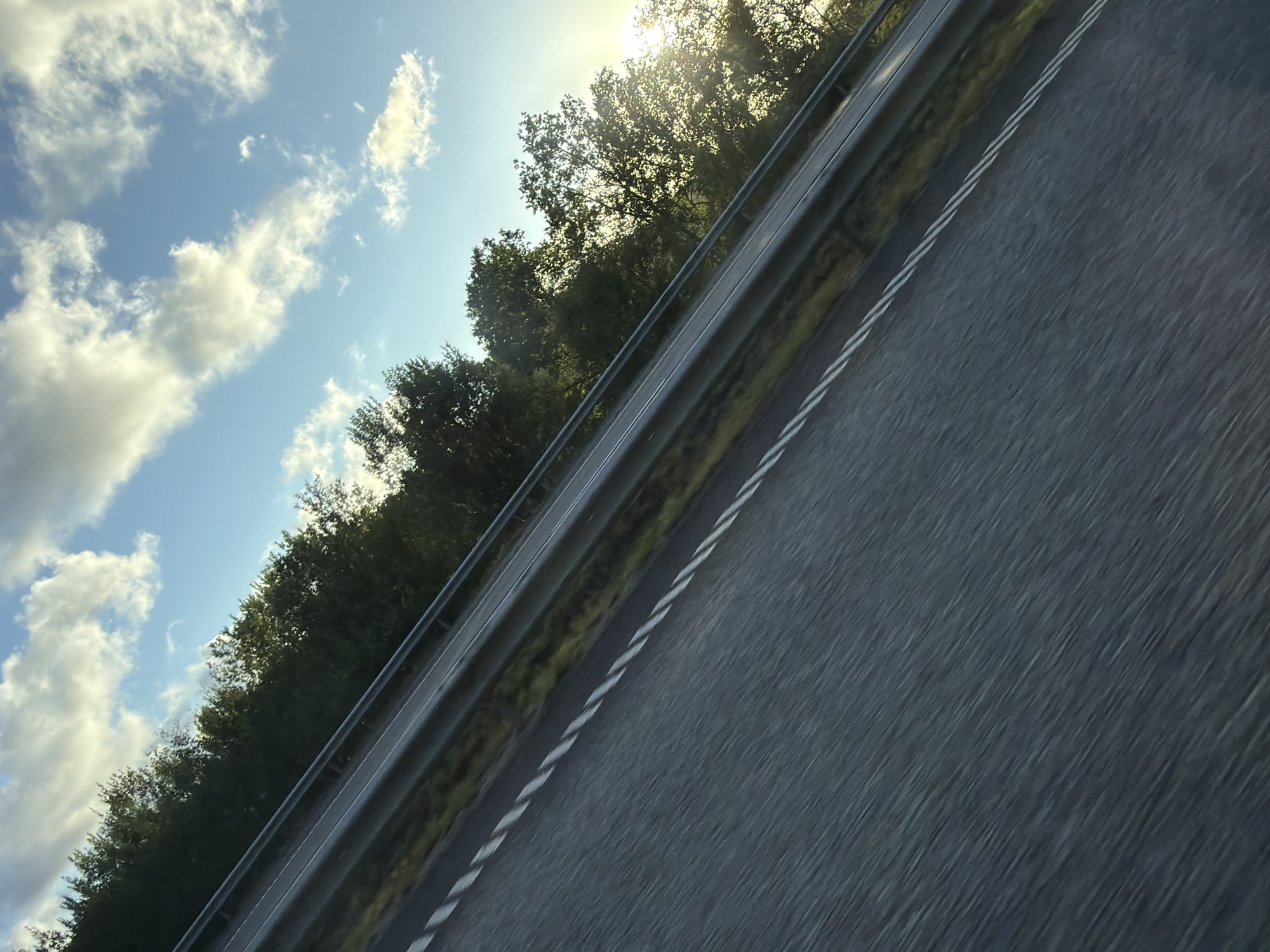 A tilted, close-up shot captures a highway's asphalt lane in Ödsmål, Sweden. The lane is marked with white dashed lines and appears blurred due to motion. A silver guardrail runs parallel to the lane, separating it from a grassy verge. Beyond the verge, a dense line of green trees flanks the road. The sky above is a bright blue, dotted with fluffy white clouds illuminated by the sun, which is peeking through the trees on the right side of the frame, casting a warm glow. The perspective suggests the photo was taken from a moving vehicle.