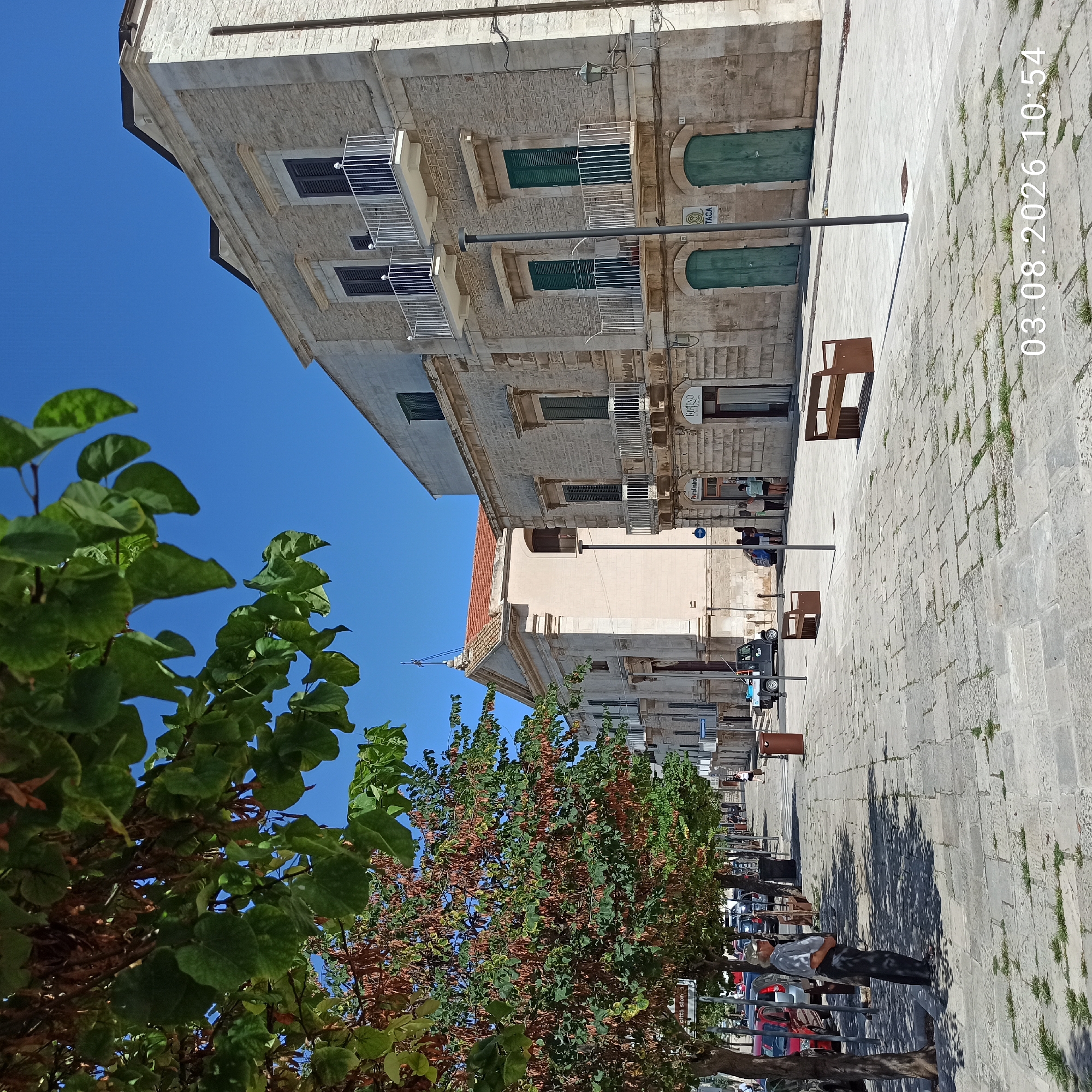 A street scene in Ruvo di Puglia, Italy, on a clear day. The image is dominated by the architecture of the buildings lining the street, which are made of pale stone. The building on the right has multiple floors with balconies and shuttered windows. To the left, a church with a red-tiled roof and a cross on top is visible. The street is paved with large stone slabs, and trees with green and brown foliage line the sidewalk on the left, casting shadows. Several parked cars are visible on the left side of the street, and a small electric utility vehicle is parked further down. A man in a light-colored shirt and dark pants stands on the sidewalk, facing away from the camera. There are also a few other pedestrians visible further down the street. Two brown park benches are placed on the paved area. The date and time "03.08.2026 10:54" are superimposed on the lower right portion of the image.