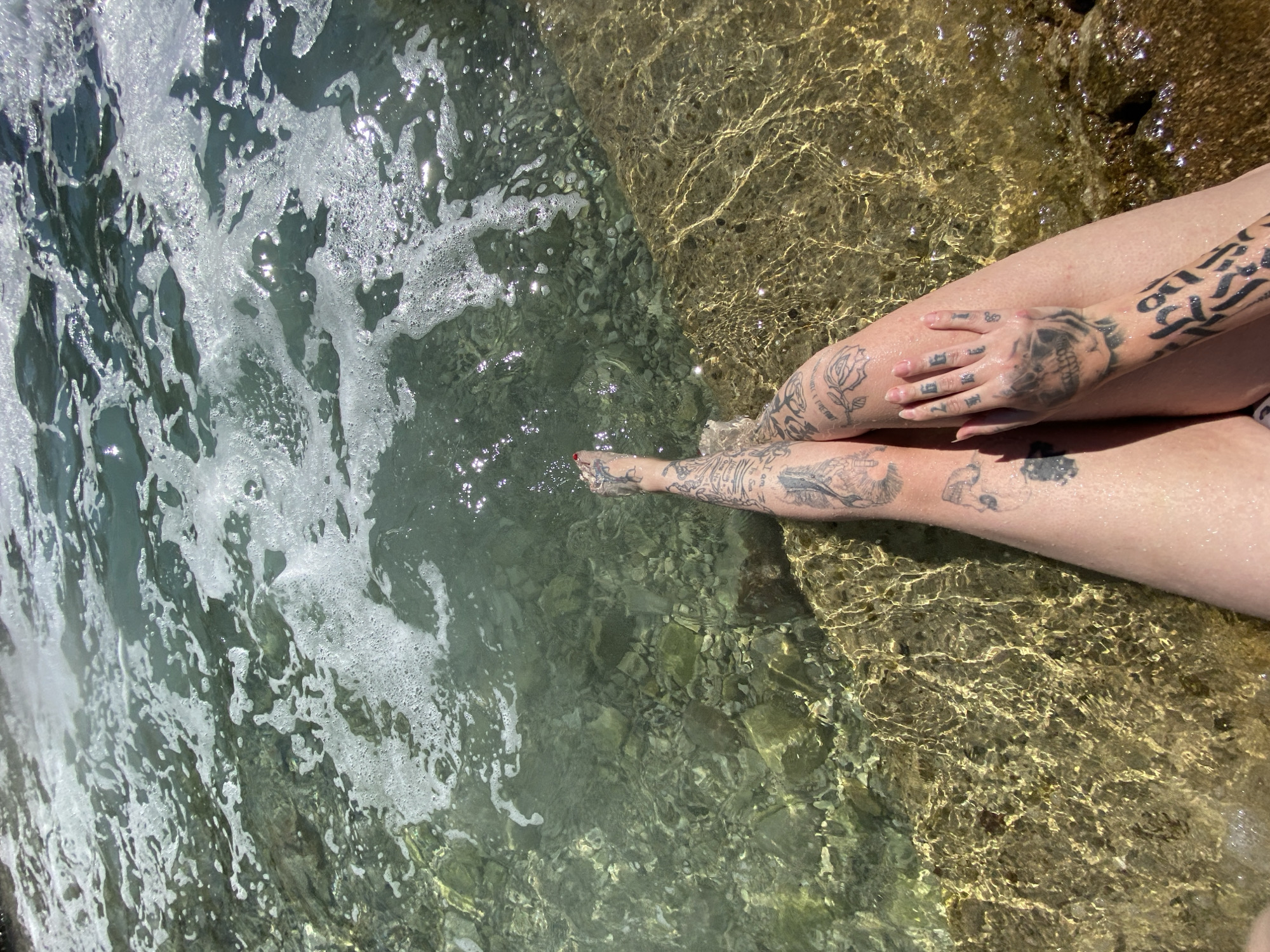 The photograph depicts a pair of crossed legs with extensive tattoos resting on a rock formation partially submerged in clear, shallow water. The water is light green in color with sunlight reflecting off its surface. The legs are light-skinned and feature tattoos on the arms, hands, legs, and feet. The tattoos appear to be of various designs, including skulls, roses, and text. The legs are positioned on a rocky surface, with the clear water around the rocks revealing small pebbles and stones at the bottom. The location is likely near the coast in Bar, Montenegro, given the rocky shoreline and clear water.