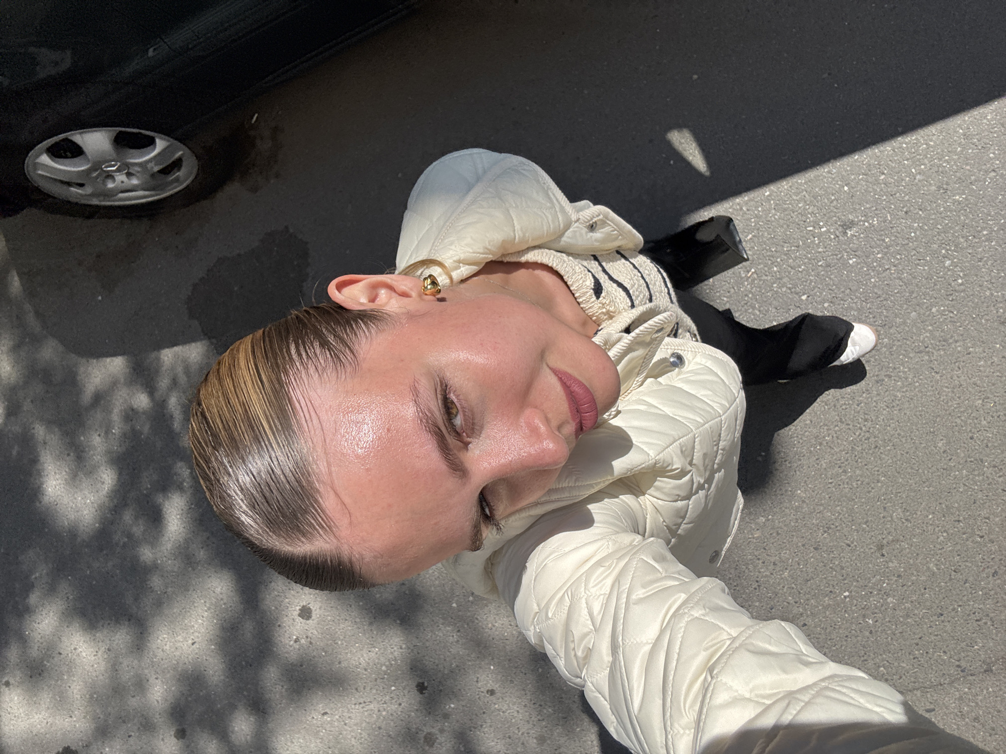 A close-up, high-angle outdoor shot captures a young woman from an elevated perspective, oriented diagonally across the frame. Her face is in a three-quarter profile, looking slightly upwards with a serene expression, a hint of a smile on her lips. Her fair skin appears dewy or subtly highlighted by the sun. She has light brown hair, slicked back, and wears natural-looking makeup with defined brows and pink lipstick. A prominent gold hoop earring adorns her right ear.
She is dressed in a cream or off-white quilted jacket, its texture clearly visible. Beneath the jacket, a striped top, possibly cream and black, is visible around her neck. Her left arm, covered by the jacket, is bent and visible in the foreground.
The setting is outdoors on a paved surface, likely asphalt or concrete, with visible textures and small debris. Strong, distinct shadows fall across the ground, indicating bright sunlight, characteristic of midday or early afternoon. In the upper left corner of the frame, part of a black car, including its tire and wheel hubcap, is visible. In the lower right, a glimpse of her white sneaker and a black bag can be seen. The overall scene suggests a casual outdoor moment in a sunny environment, possibly on a street or parking area in Tbilisi, Georgia. There is no visible text in the image.