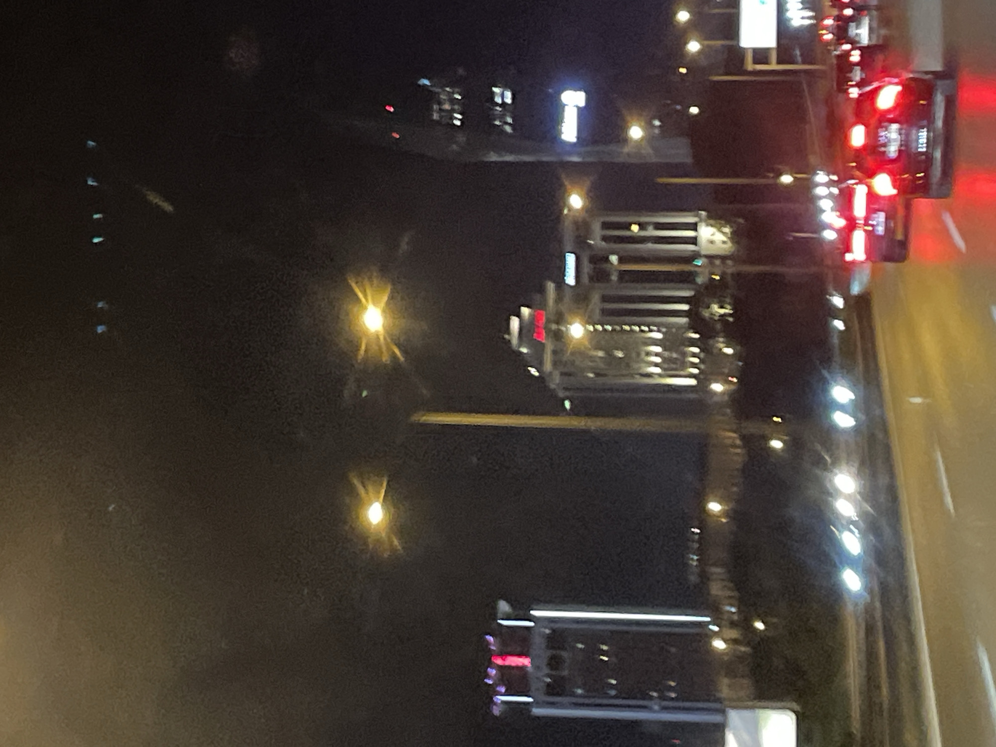The image depicts a nighttime scene of a highway in Ataşehir, Turkey, with several illuminated buildings in the background. The focus is on the road where multiple cars are driving, their headlights and taillights creating streaks of light. The buildings in the background are modern high-rises, some of which are lit up with architectural lighting. One prominent building on the right has a sign that reads "Allianz". The sky is dark, suggesting it is late at night. The overall impression is one of urban activity and movement on a highway at night.
