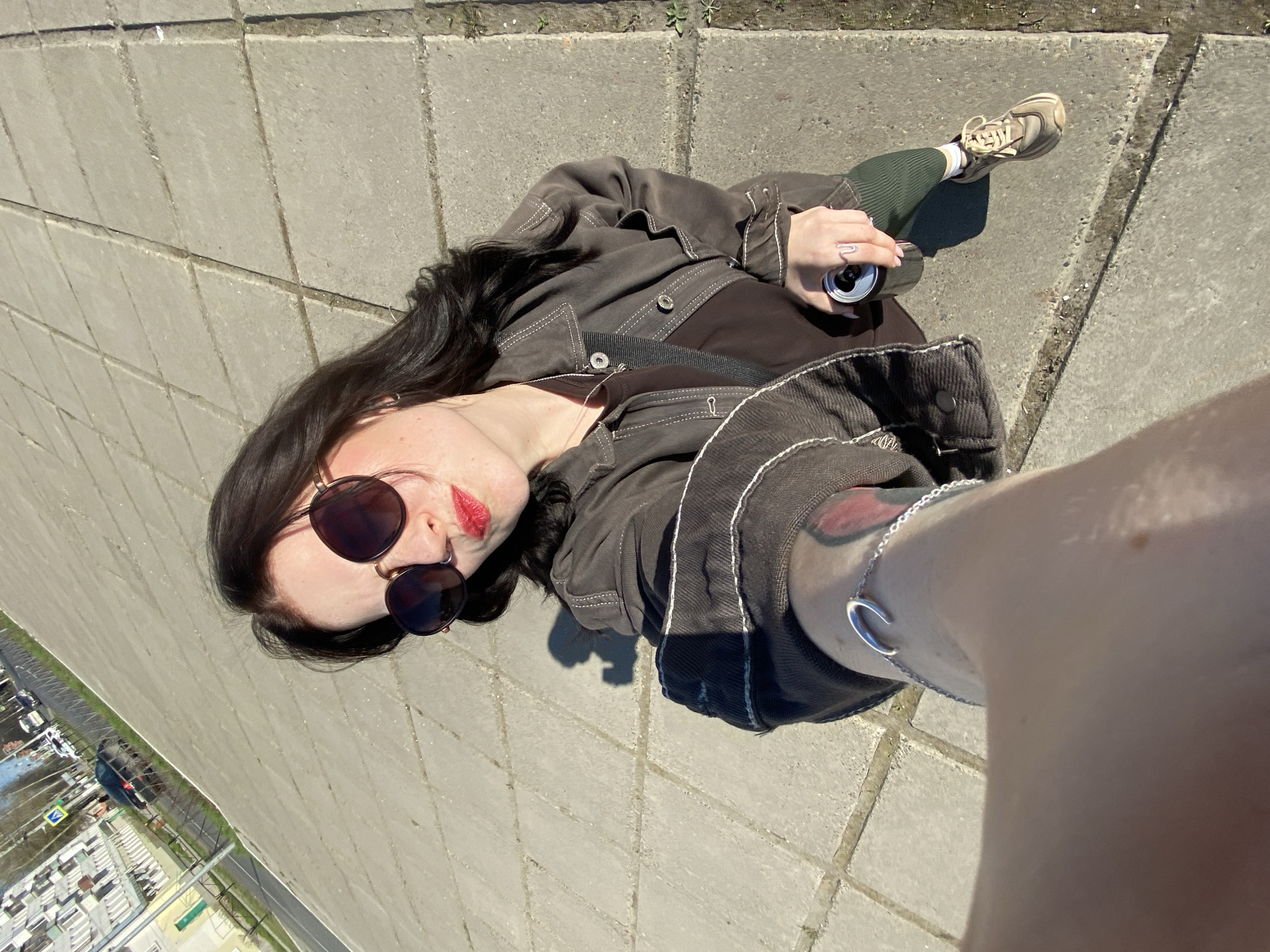 A woman is lying on a gray tiled surface, likely a pedestrian walkway. She wears sunglasses and red lipstick. Her dark hair contrasts with her fair skin. She is dressed in a dark jacket and holds an open beverage can. A leg clad in a green sock and a sneaker is visible near the can. A silver bracelet adorns her wrist. In the background, a cityscape is visible through a green fence. Based on the surroundings, the location is Chaikovsky, Russia.