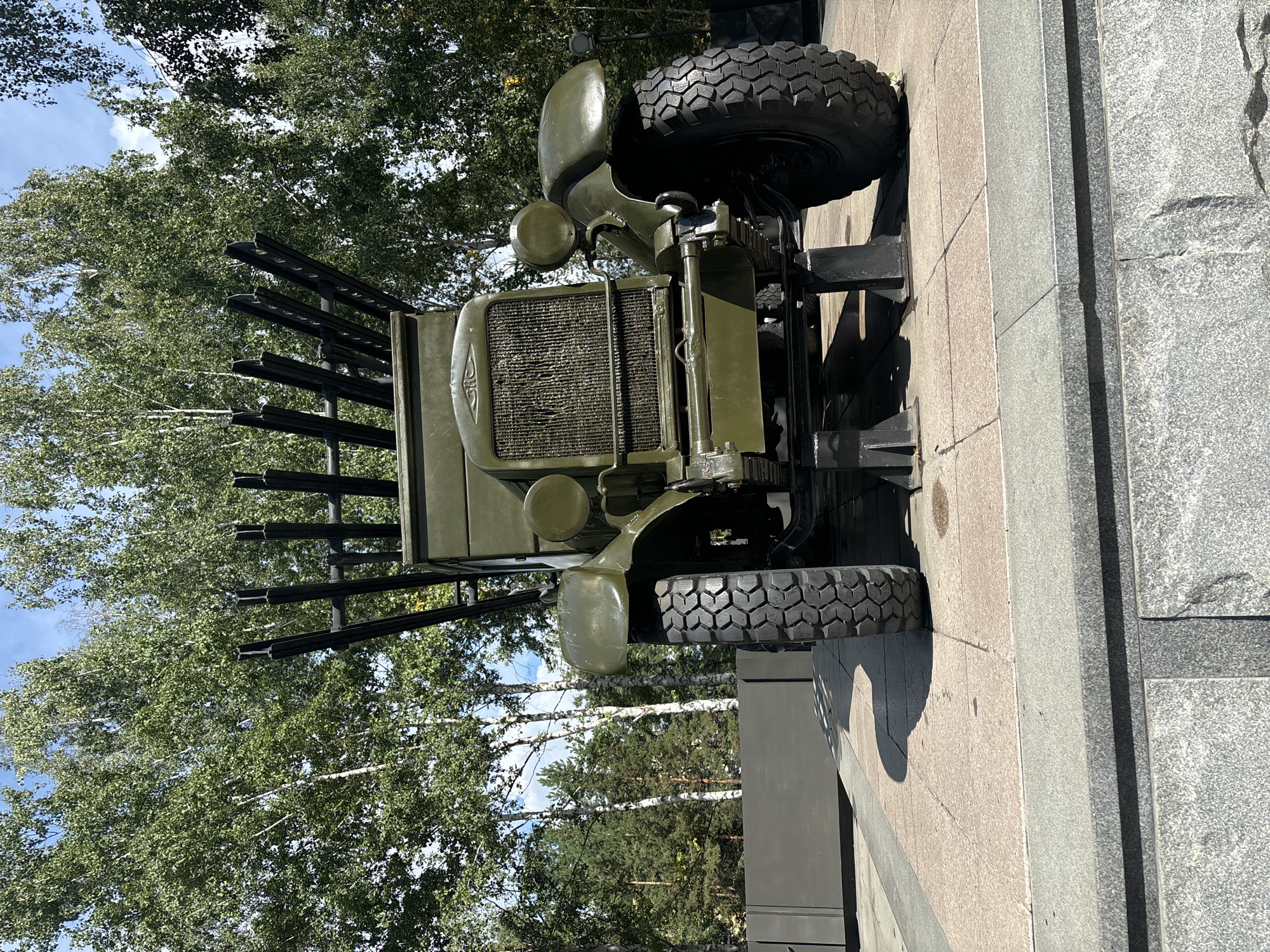 An outdoor, eye-level, full shot depicts a vintage Soviet BM-13 Katyusha rocket launcher truck, painted in olive drab, positioned on a paved display area. The truck is the central subject, with its distinctive rocket tubes angled upwards from the rear bed. The vehicle features large, rugged tires and a prominent front grille. The display area is surrounded by granite and stone structures. In the background, a dense forest of birch and pine trees creates a natural backdrop under a bright, partly cloudy sky. The scene is devoid of any people. This display is located at the Glory Monument in Novosibirsk, Russia.