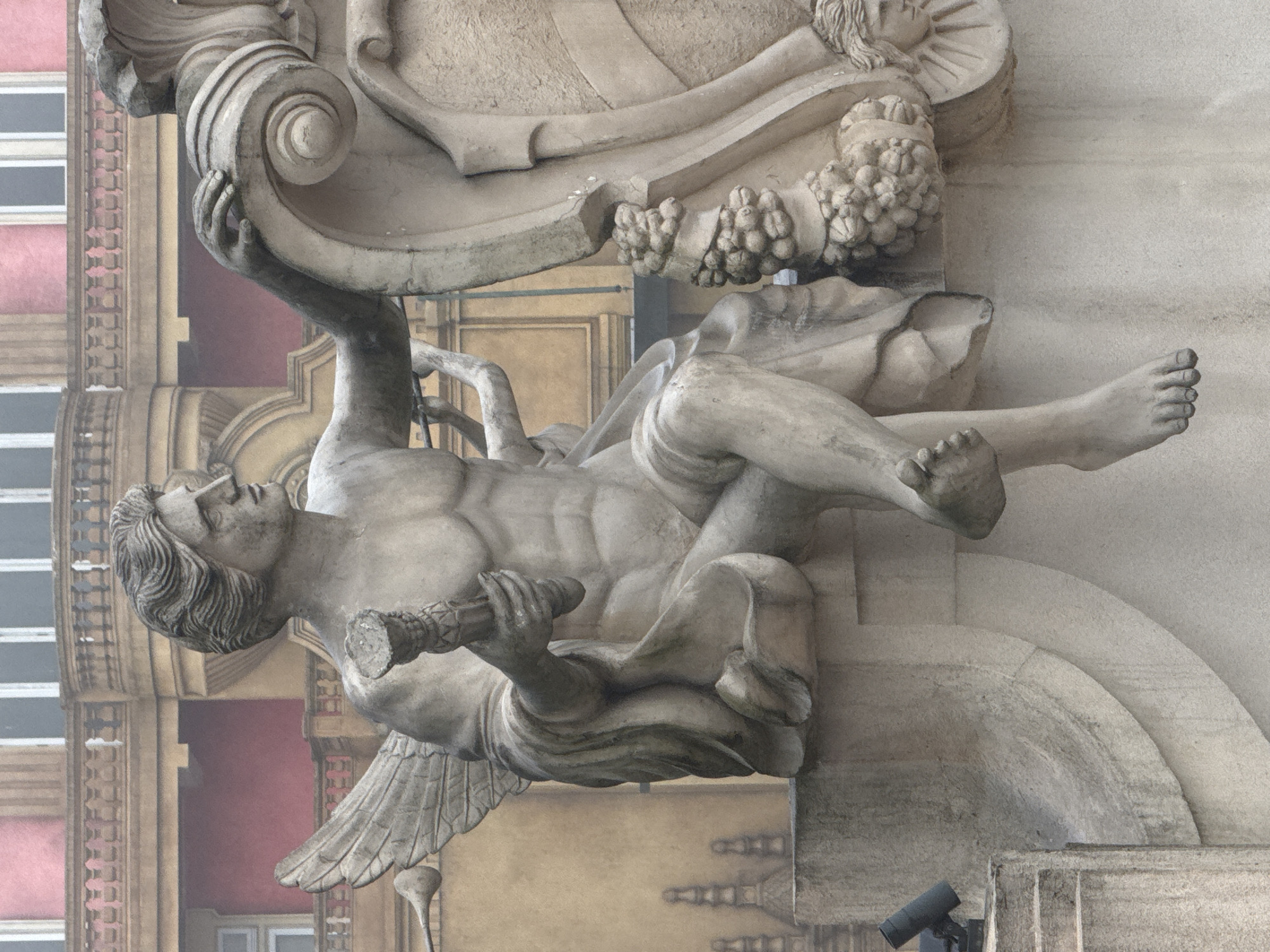 The image depicts a weathered stone sculpture of a winged male figure supporting an ornate architectural element. The figure is reclining, holding a cylindrical object in one hand, and appears to be straining under the weight of the structure above. The architectural element includes a shield-like shape adorned with carvings, potentially grapes or berries. The backdrop shows a building with windows and decorative facade elements, possibly in a European style. A black security camera is mounted on a wall at the bottom of the image. The location in Đà Nẵng, Vietnam is not evident from the image content.