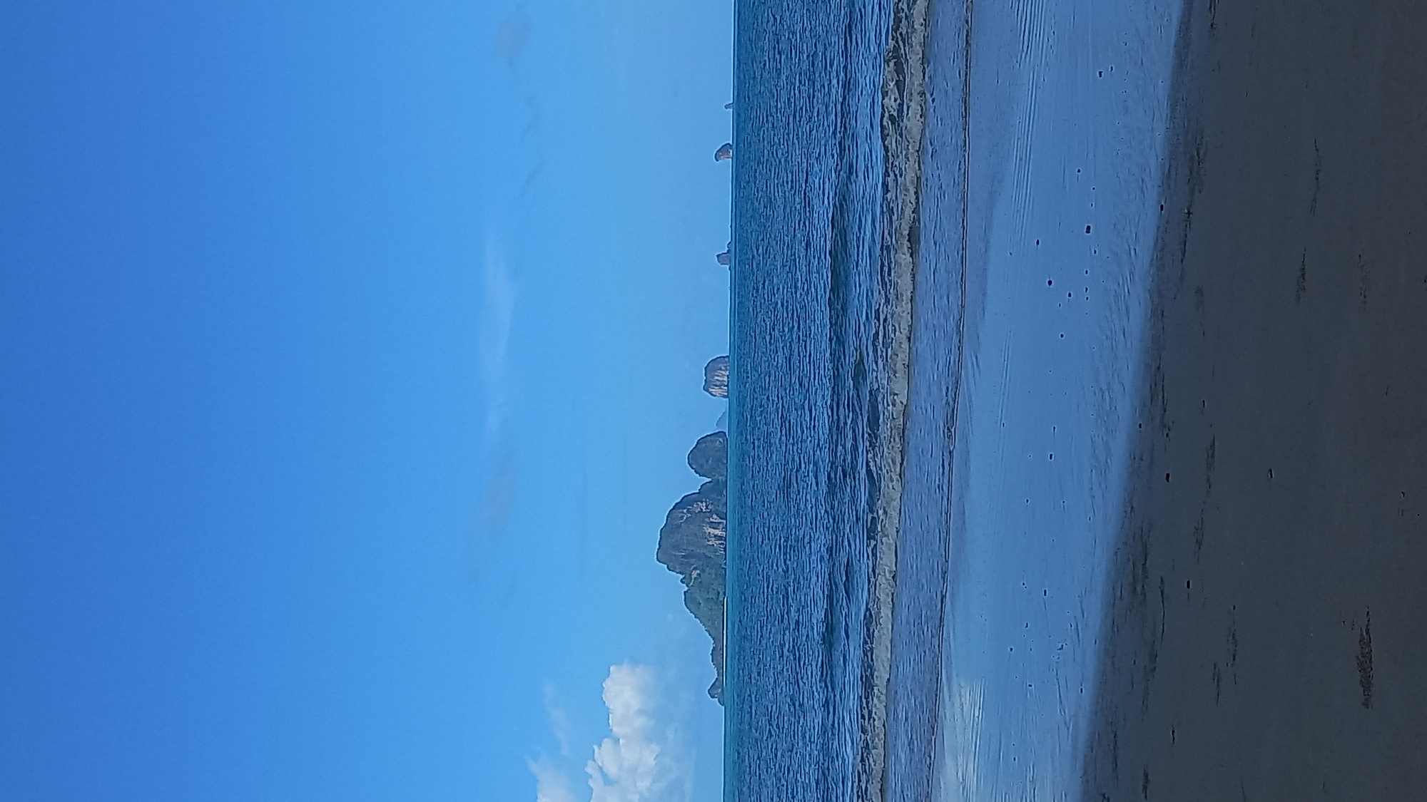 The image shows a beach scene in Ban Klong Haeng, Thailand. The scene includes a sandy beach, a shoreline with gentle waves, and several limestone rock formations in the distance. The sky is clear blue with a few clouds. No people are visible.
