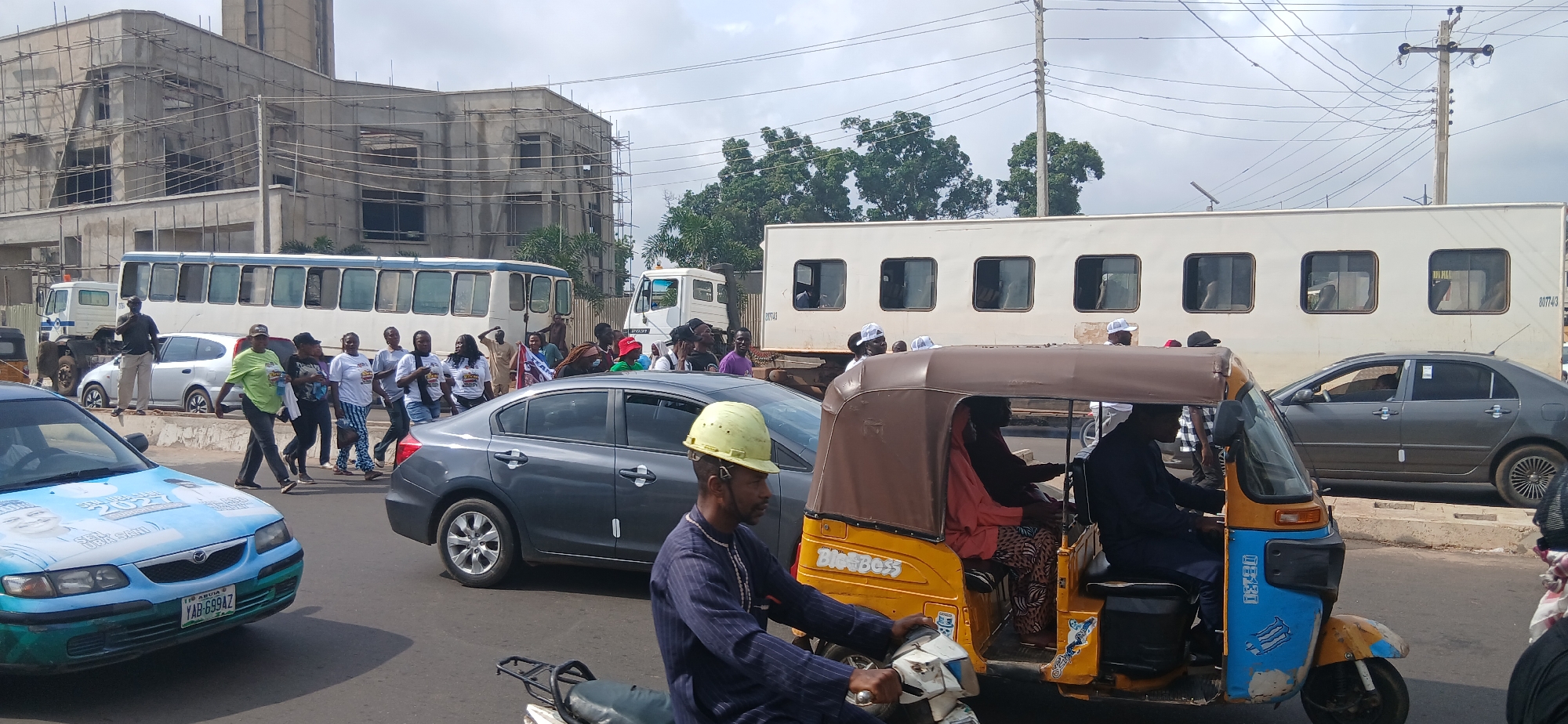 In Unguwar Shanu, Nigeria, a street scene unfolds with multiple vehicles and pedestrians. A light blue Mazda sedan, adorned with political campaign imagery and the registration plate YAB-699AZ, is prominent in the foreground. To its right, a dark gray sedan is partially visible. A yellow and brown auto-rickshaw, a common mode of transport, is in the mid-ground, carrying passengers. Behind the auto-rickshaw, a long, white trailer with multiple square windows is stationary, possibly parked. Further back, a white bus with many windows is also parked. A construction site with scaffolding and unfinished buildings is visible on the left. A group of people, some wearing white t-shirts with logos, are walking across the street. The sky is overcast with visible power lines. A man wearing a dark blue robe and a light green hard hat is riding a motorcycle in the foreground, to the left of the auto-rickshaw.