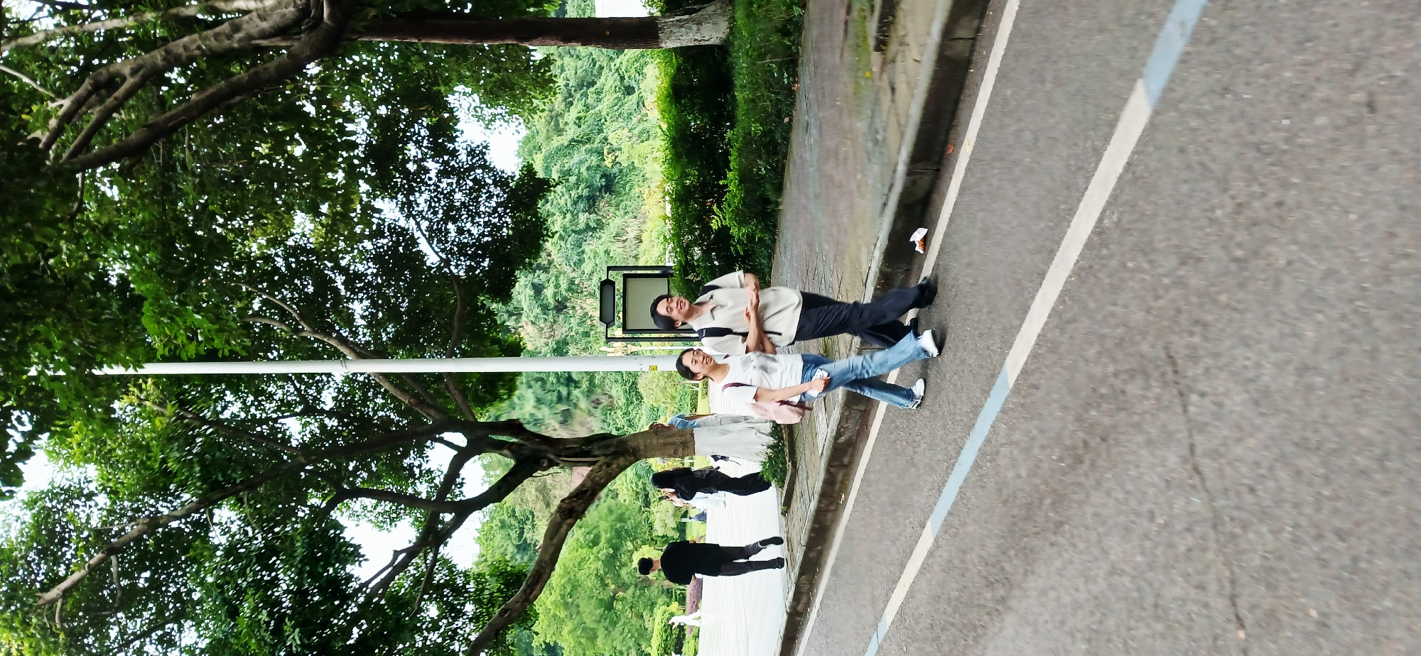The image captures an outdoor scene in Chongqing High-tech Zone, China. A group of individuals are present, with two people standing closer to the camera. A male, wearing a light-colored shirt and dark pants, is standing behind a female who is wearing a white t-shirt and jeans. They are standing on a paved area, with a road visible on the right side. The road has a white lane marker. A tree with dense foliage is prominent on the left side of the frame. In the background, lush greenery and other trees can be seen. Other people are walking behind the couple on a concrete path.