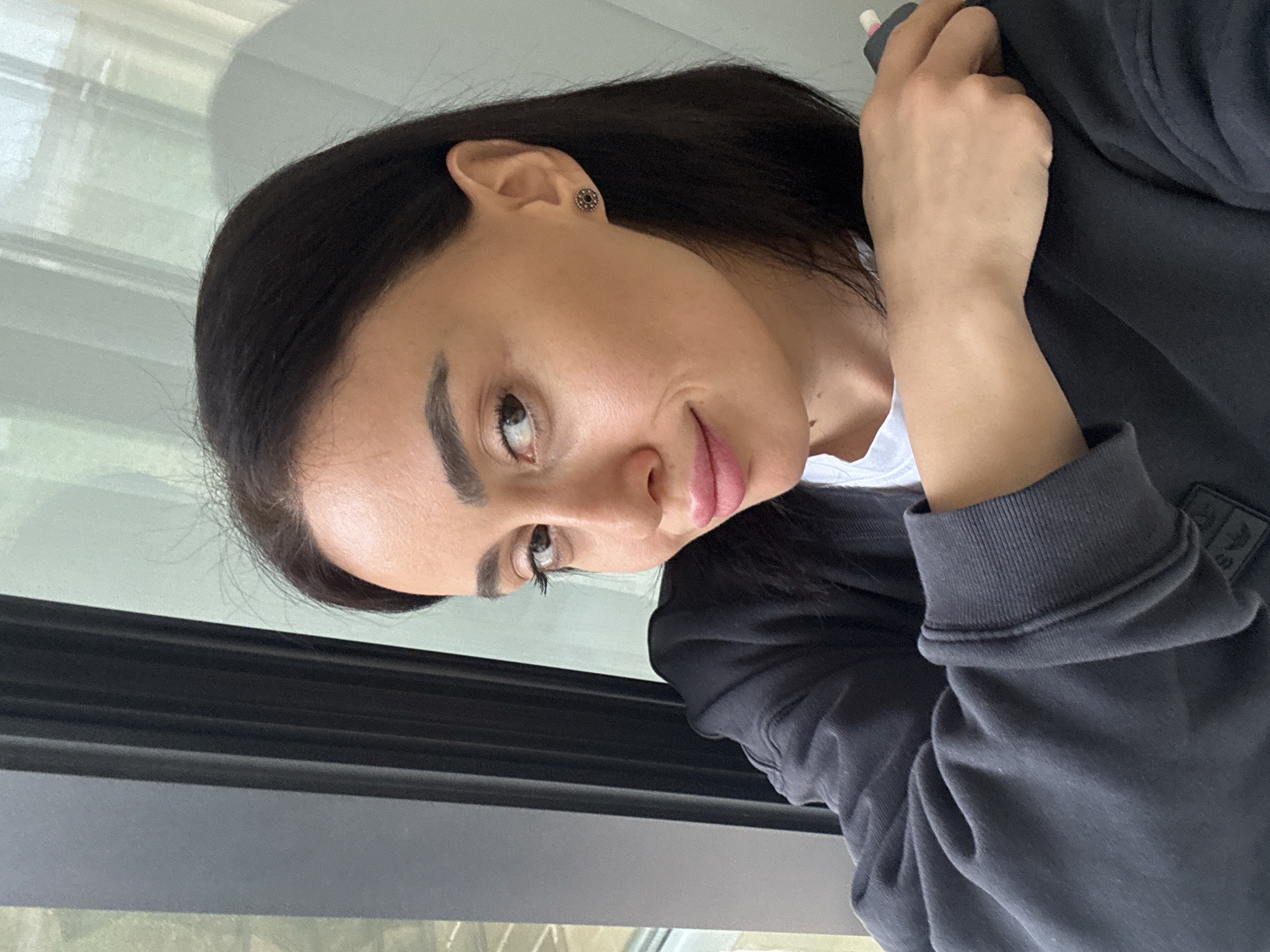 A female subject is depicted in a close-up, indoor portrait. She has dark brown hair, brown eyes, well-defined eyebrows, and is wearing pink lipstick, exhibiting a faint smile. A circular, dark-toned stud earring is visible in her right ear. She is dressed in a dark grey or black long-sleeved garment, possibly a sweatshirt, with a white collar visible beneath. Her right arm is raised, with her right hand resting on her left shoulder, holding a small white object with a pink tip, consistent with a pen or pencil with an eraser. The background consists of a large window to the subject's left, revealing blurred green foliage and indistinct structures outdoors. To her right, a light-colored wall or curtain with faint vertical textures is visible. The lighting is soft and appears natural. The specific location within Tbilisi, Georgia, cannot be determined from the visual information provided in the image.