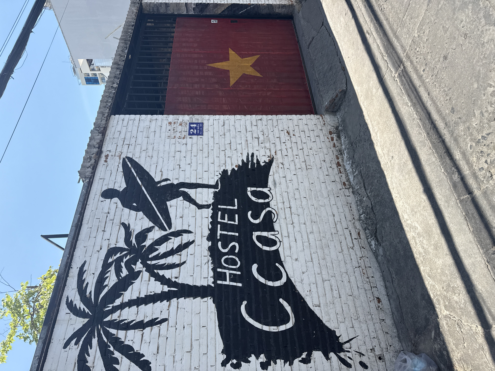 The image shows a white brick wall with a mural of a surfer silhouette against palm trees. The text "HOSTEL CASA" is painted vertically in white letters on the mural. Above the mural, a red garage door with a yellow star, the flag of Vietnam, is visible. A blue street sign with the number "24" and Vietnamese text is also present on the wall. The scene appears to be outdoors, likely on a sunny day, given the bright lighting and strong shadows. The setting is urban, with a hint of greenery from a nearby tree and power lines overhead. The overall impression is of a casual and inviting establishment, possibly a hostel in a coastal area given the surf imagery. The style of the mural suggests a relaxed, possibly tropical atmosphere.