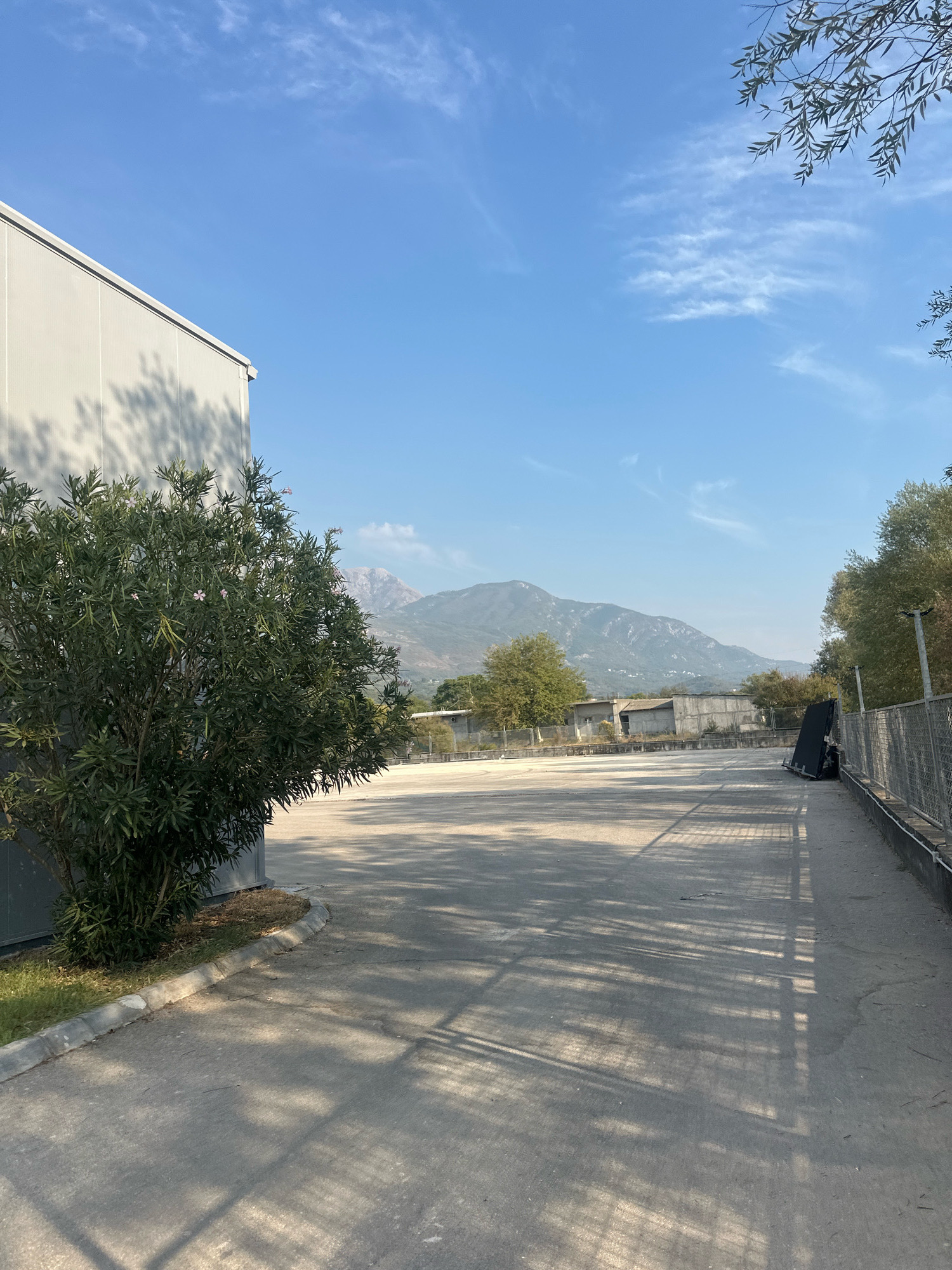 A wide, paved lot is bordered by a building on the left and a chain-link fence on the right. In the background, mountains rise under a clear blue sky with scattered wispy clouds. A large, green shrub with small pink flowers is positioned in the foreground to the left of the lot. The pavement is mostly bare, with shadows from the surrounding trees creating a dappled pattern. Some scattered debris is visible on the ground, including a dark, upright object leaning against the fence on the right side of the lot. The scene appears to be in an industrial or semi-developed area, with some low-lying structures visible in the middle ground. The location is identified as Radanovići, Montenegro.