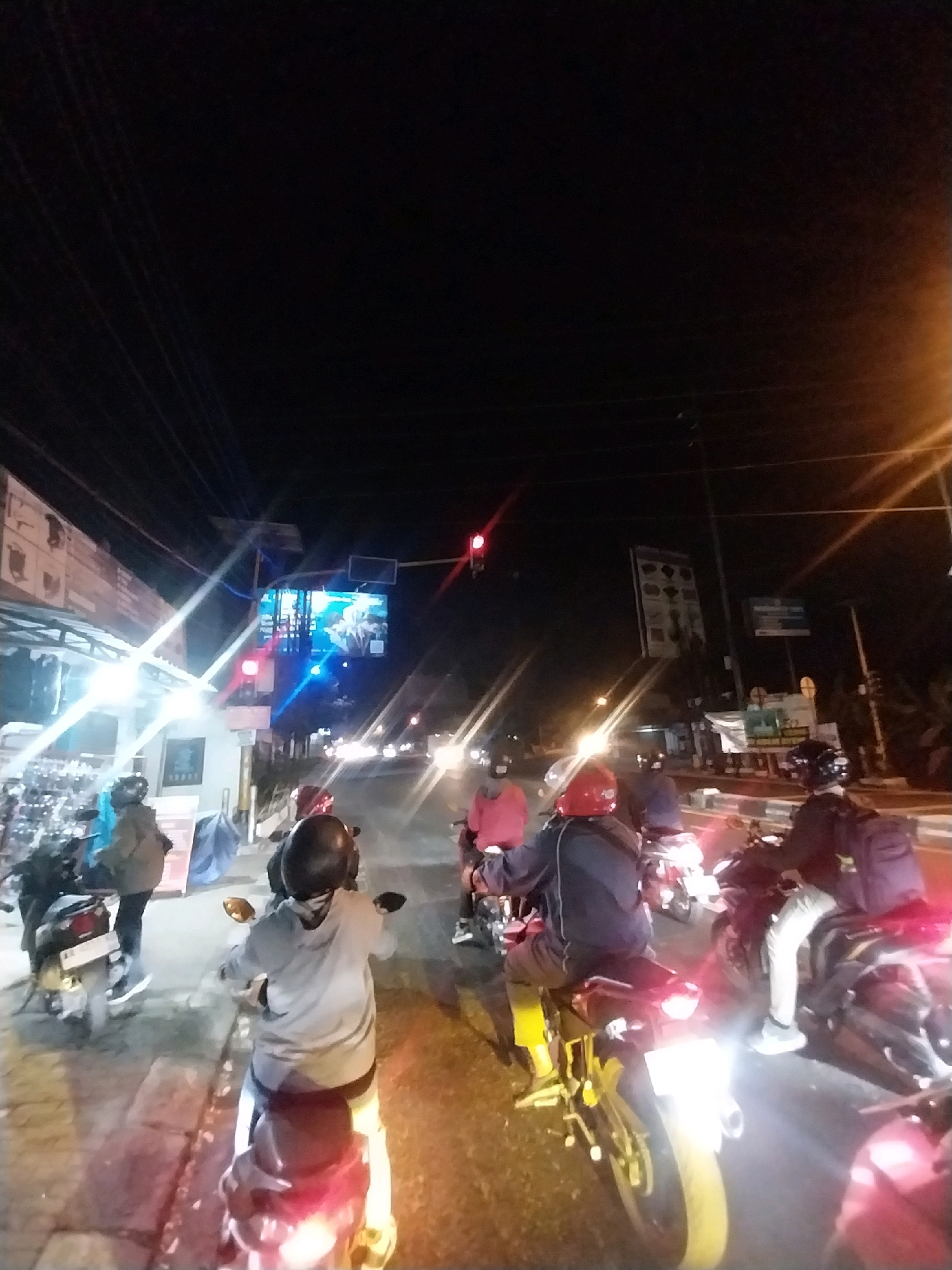 A group of motorcyclists are stopped at a traffic light on a dark, paved road at night, likely in Tempel, Indonesia. The foreground features several individuals on motorcycles, illuminated by streetlights and the headlights of their own vehicles. One person in the center foreground wears a grey hoodie and a dark helmet, sitting on a yellow motorcycle. To their left, another motorcyclist in a blue jacket and a blue helmet is partially visible. Further back, a person in a pink jacket rides a motorcycle. To the right, a motorcyclist in a dark jacket and a dark helmet with a purple backpack is stopped. In the background, buildings with illuminated signs are visible on the left side of the road. A large, illuminated billboard displays a moving image. A traffic light is red. The road is lined with trees and poles with electrical wires. The overall scene depicts a common urban nighttime traffic situation with multiple vehicles and pedestrians.