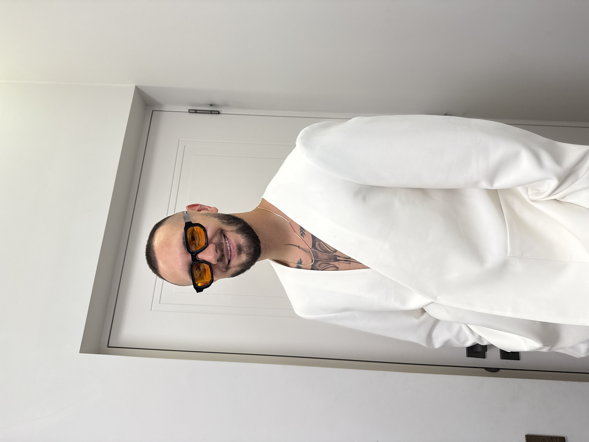 A man with a shaved head, beard, and tattoos on his chest and neck is wearing sunglasses and a white suit. He is standing in front of a white door. The setting appears to be indoors. There is no information to suggest the image was taken in Yerevan, Armenia.